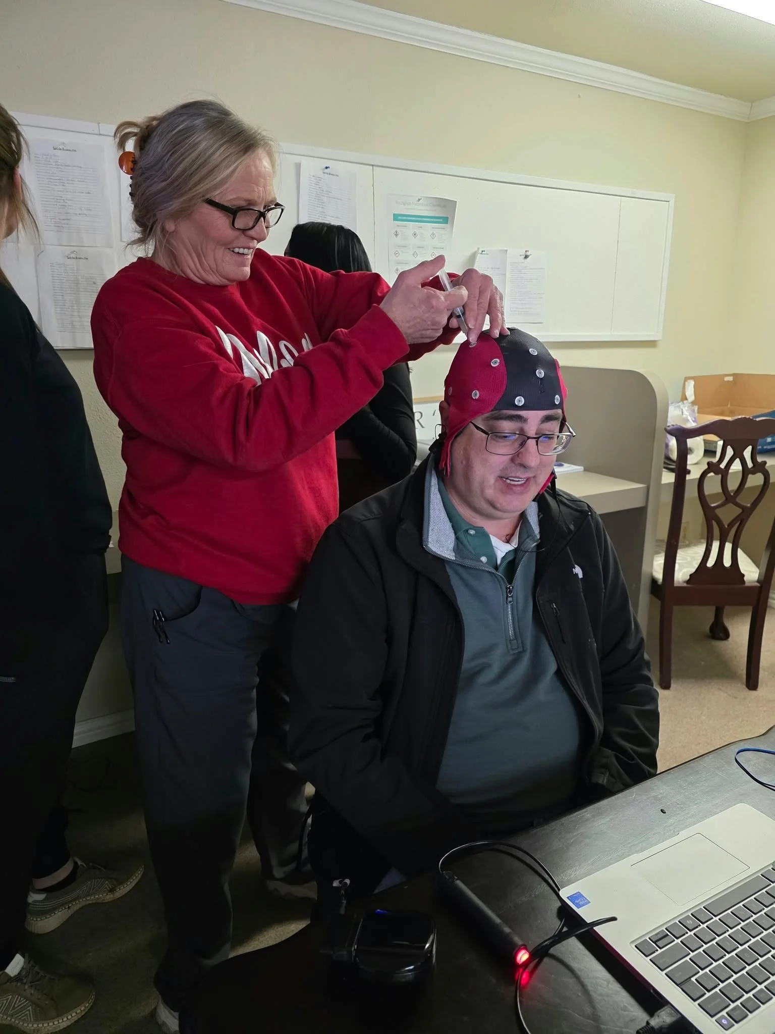 Woman adjusts a sensor-covered cap on a seated man's head during a brain activity recording session.