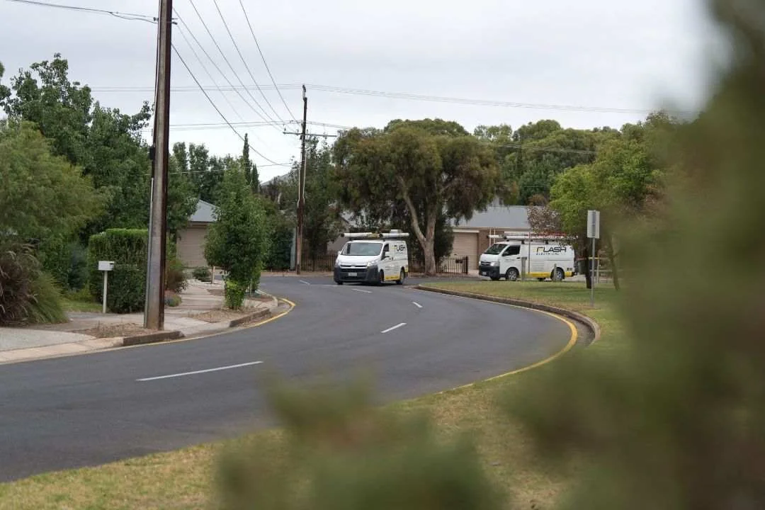 Two white 'Flash Electrical' vans driving on a curved suburban road lined with trees, houses, and utility poles, viewed through out-of-focus green foliage in the foreground.