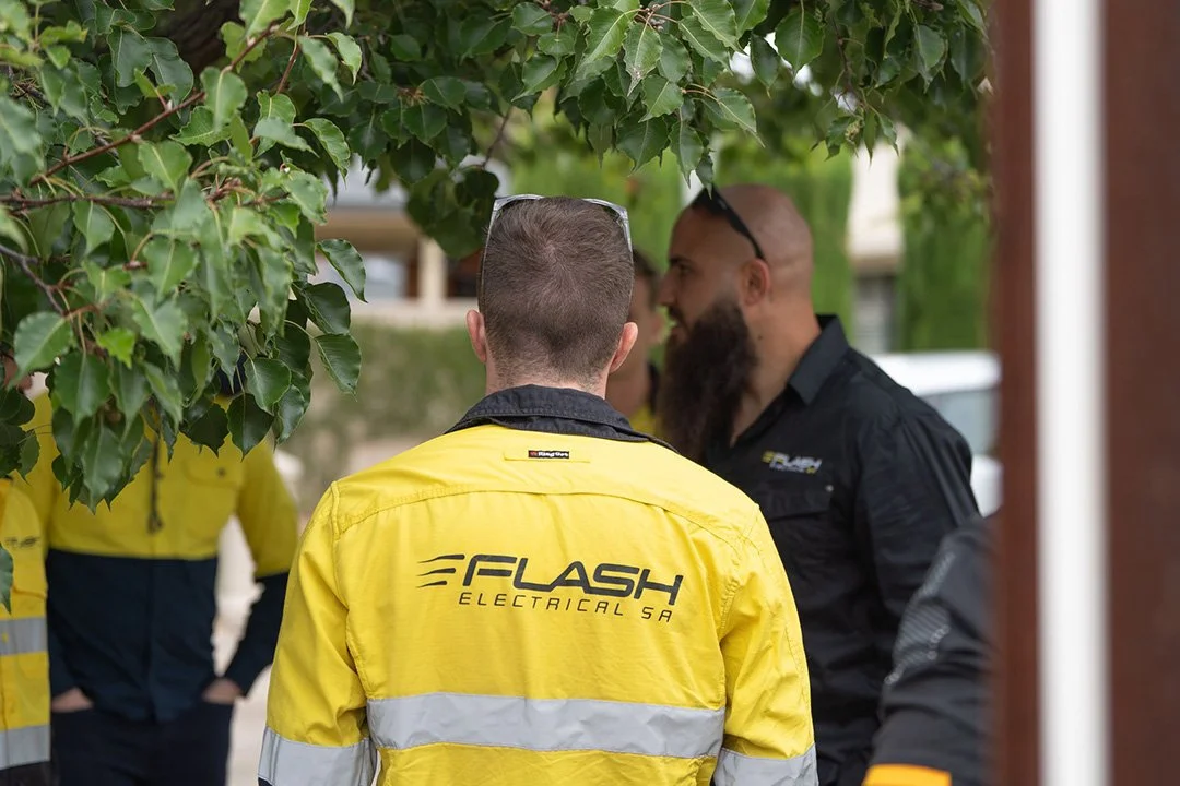 A worker viewed from behind wearing a bright yellow high-visibility jacket with the logo 'FLASH ELECTRICAL SA