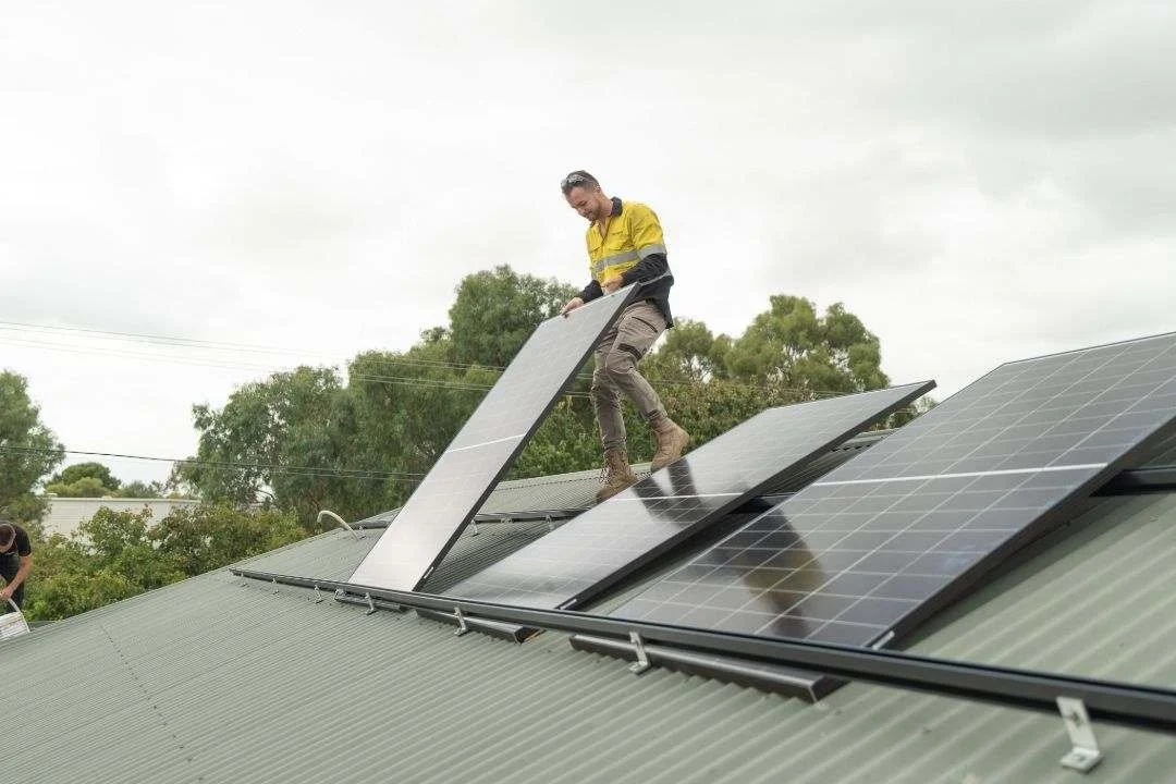 A construction worker wearing a high-visibility yellow shirt, tan work pants, and work boots stands on a corrugated metal roof,