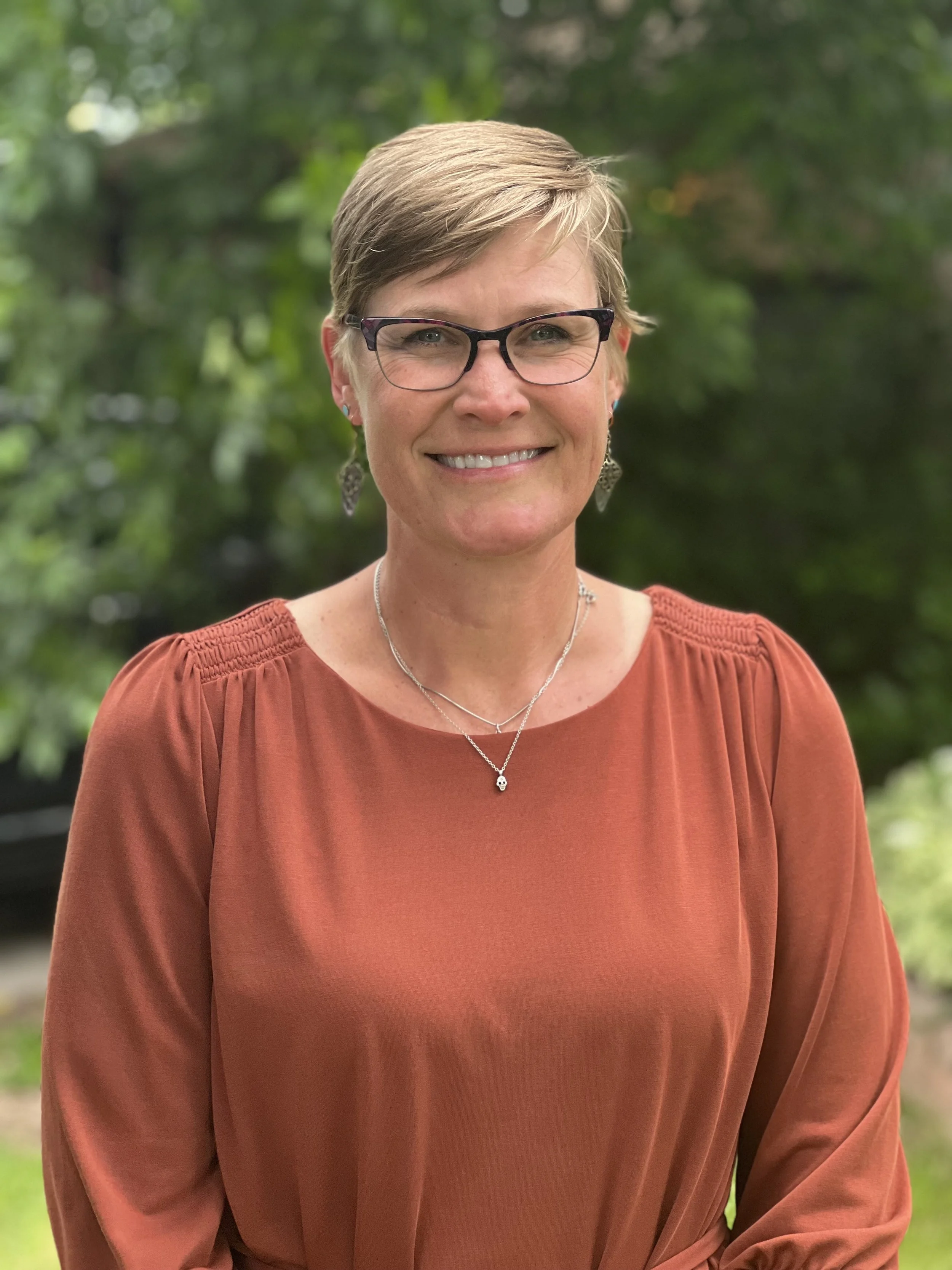 A middle-aged woman with short light brown hair, wearing glasses, earrings, a necklace, and a burnt orange top, smiling outdoors with a green, leafy background.