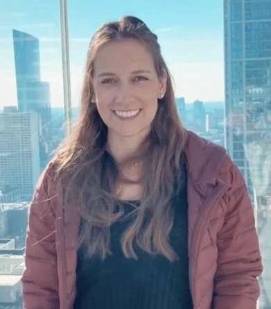 A woman smiling with long hair, wearing a maroon jacket over a black top, standing in front of a large window in a city skyscraper, with tall buildings visible in the background.
