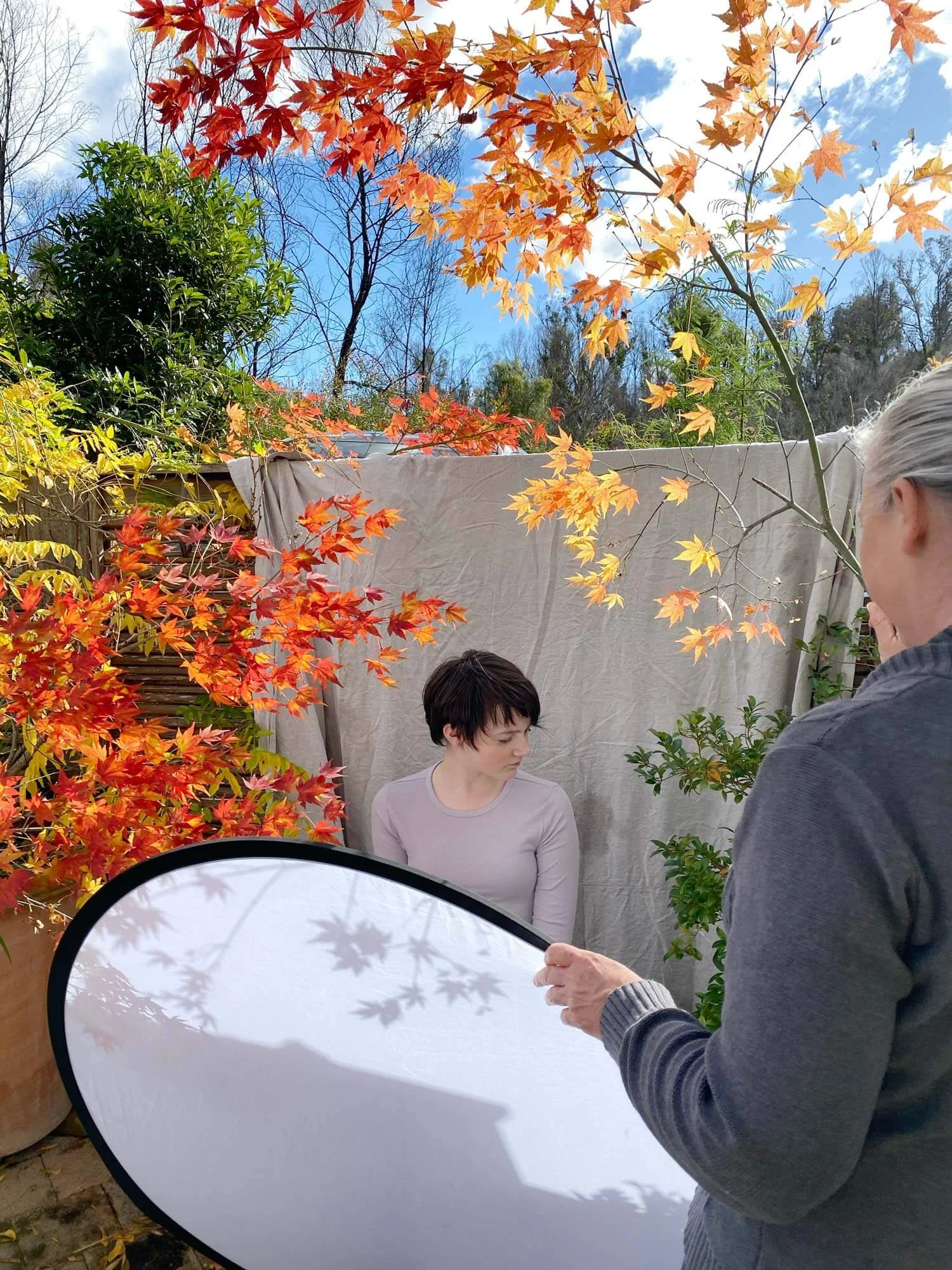 A behind the scenes of a photography shoot with photographer holding a lighting board in the foreground and with a young model next to bright orange autumn leaves in the background.
