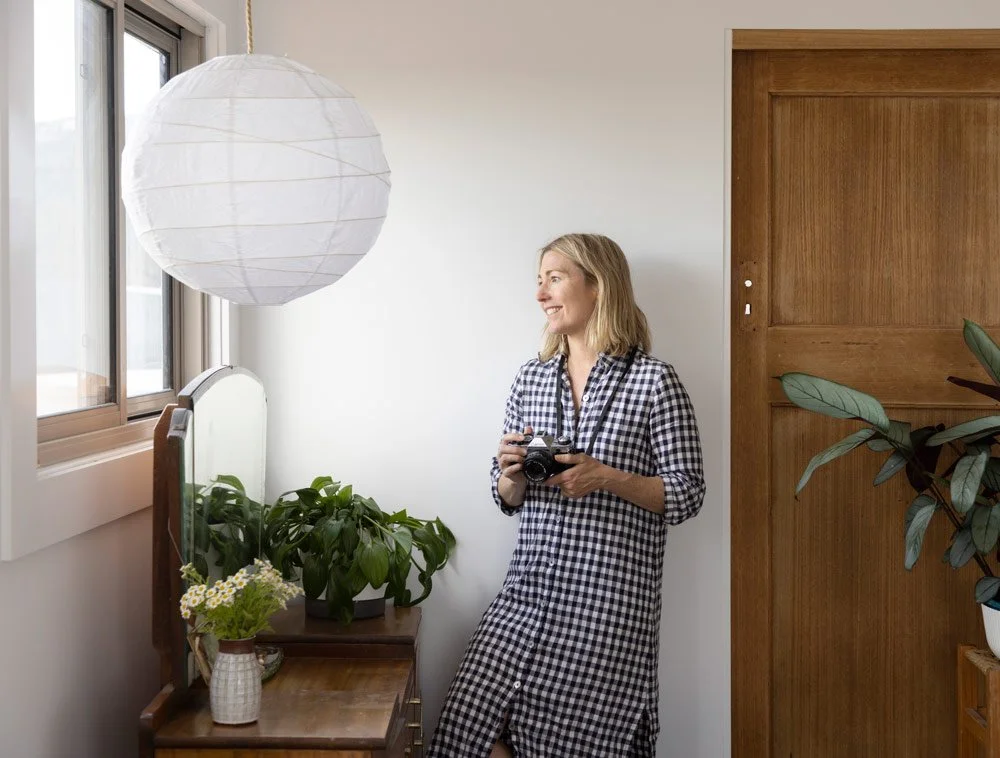 Personal brand photographer Honey Atkinson standing in a white bedroom with a film camera, surrounded by plants and natural light, showing her personal style and personality