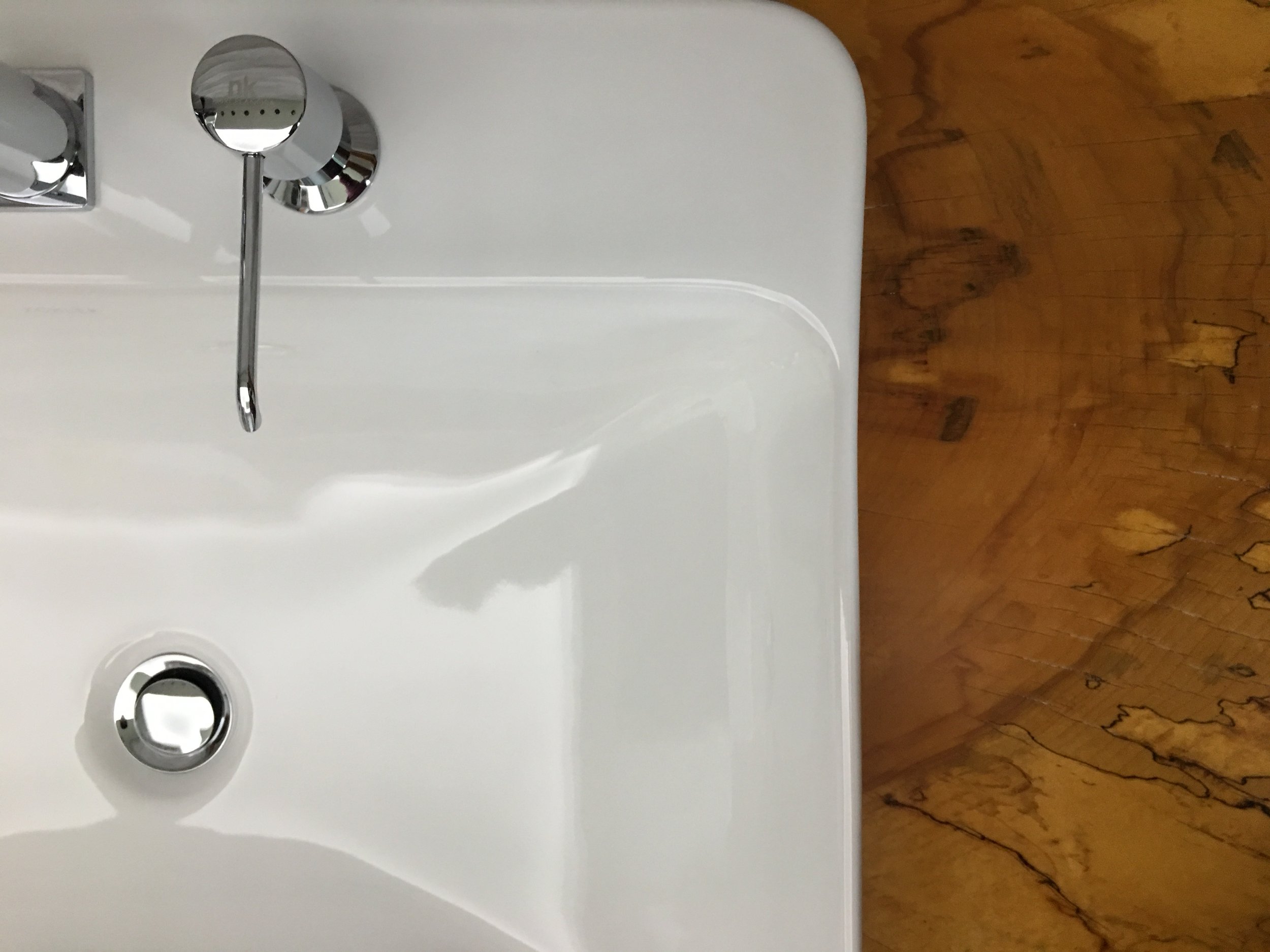 Close-up of a white sink with a chrome faucet and a drain, with a wooden floor in the background.
