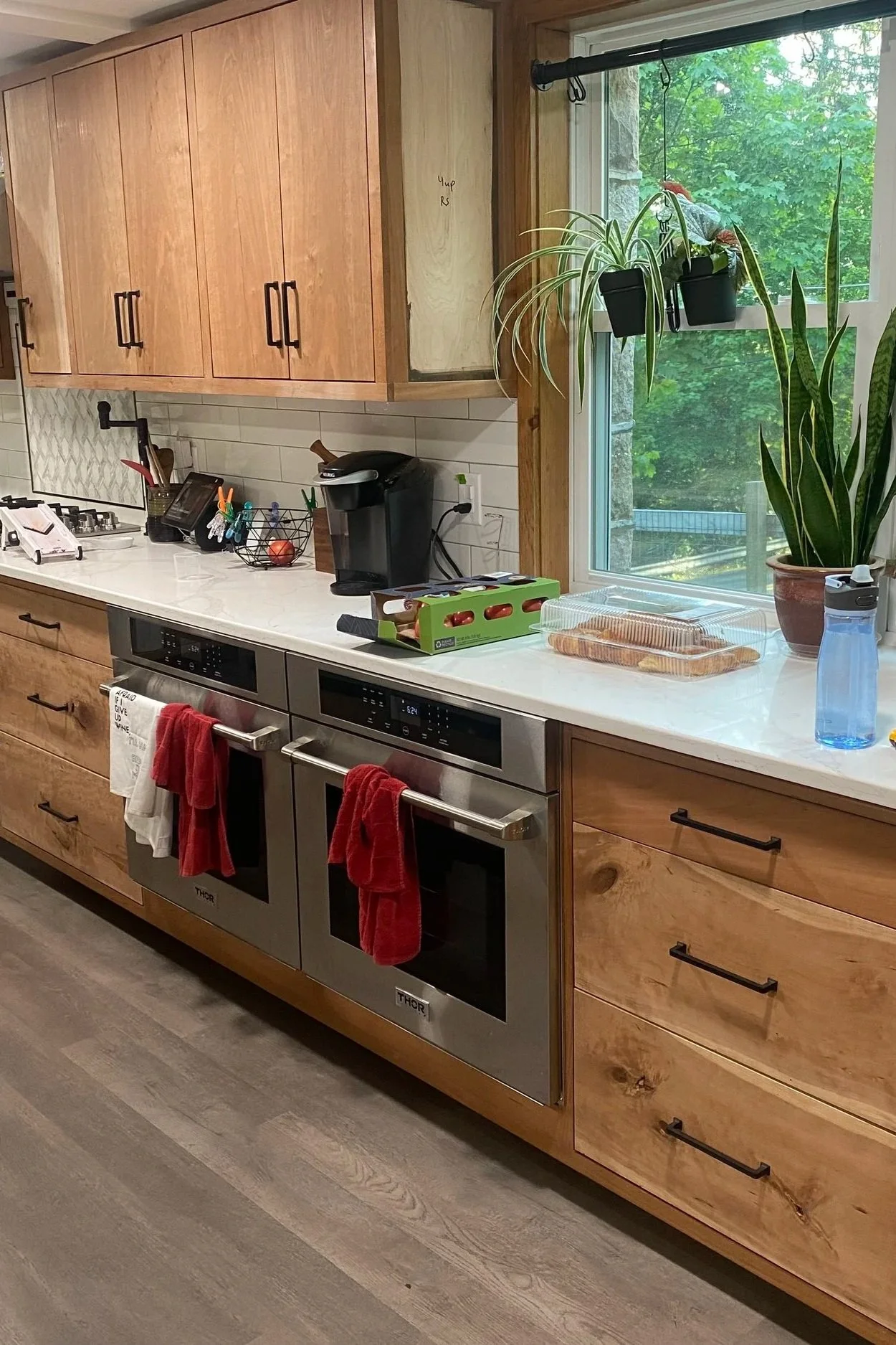Kitchen with wooden cabinets, a white countertop, a window with green trees outside, and various kitchen appliances and plants.