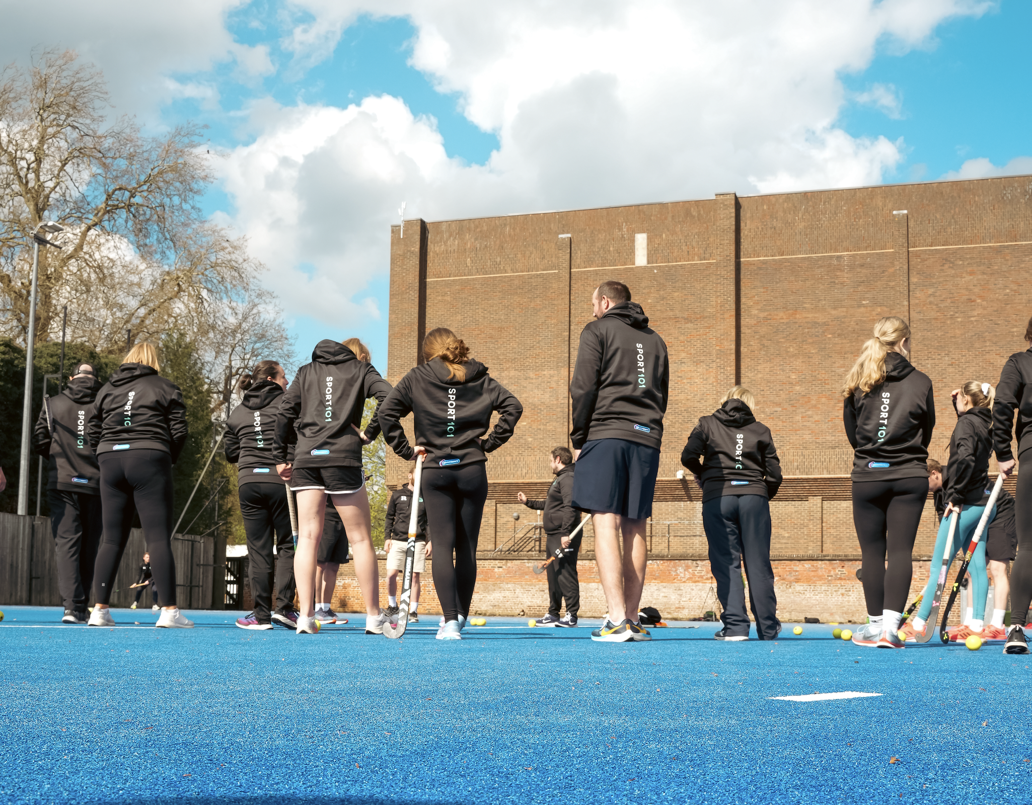 A group of people dressed in black sportswear gathered outdoors on a bright day, with some holding hockey sticks and yellow hockey balls, on a blue sports field with a brick building and leafless trees in the background.