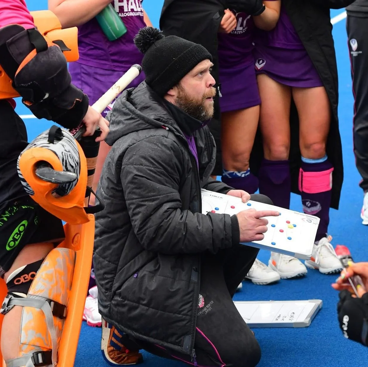 A male hockey coach kneeling on a blue ice rink, holding a white tactical board with yellow, red, and blue pegs, giving instructions to female players. The coach is wearing a black jacket and a knit cap, and the players are in purple uniforms with knee-high socks, some with pink or blue accessories. One player has a roller skate around her neck.
