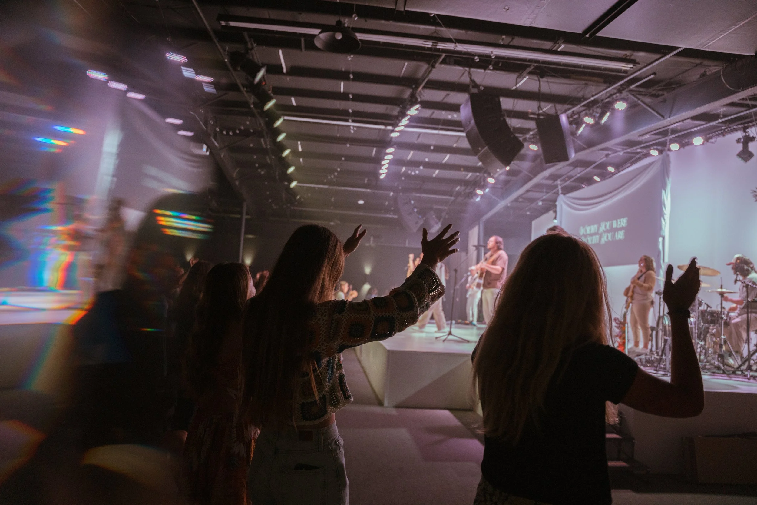 Worship event with people raising their hands in front of a stage where musicians are. Stage lights and speakers are visible overhead.