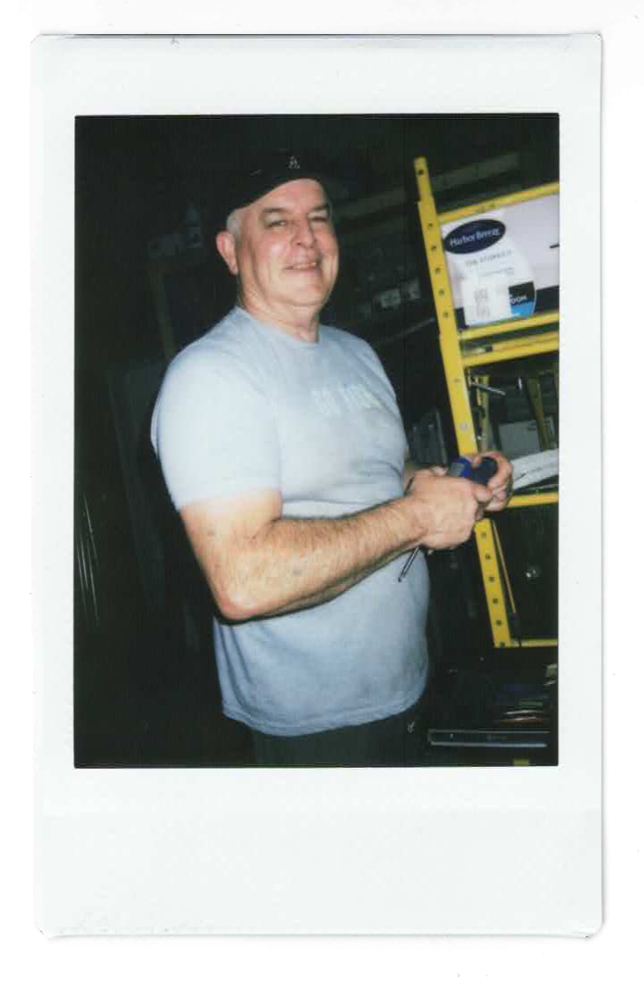 A man smiling and looking at the camera while holding a small device, standing in front of a shelf with various items, including a yellow level, in an indoor setting.