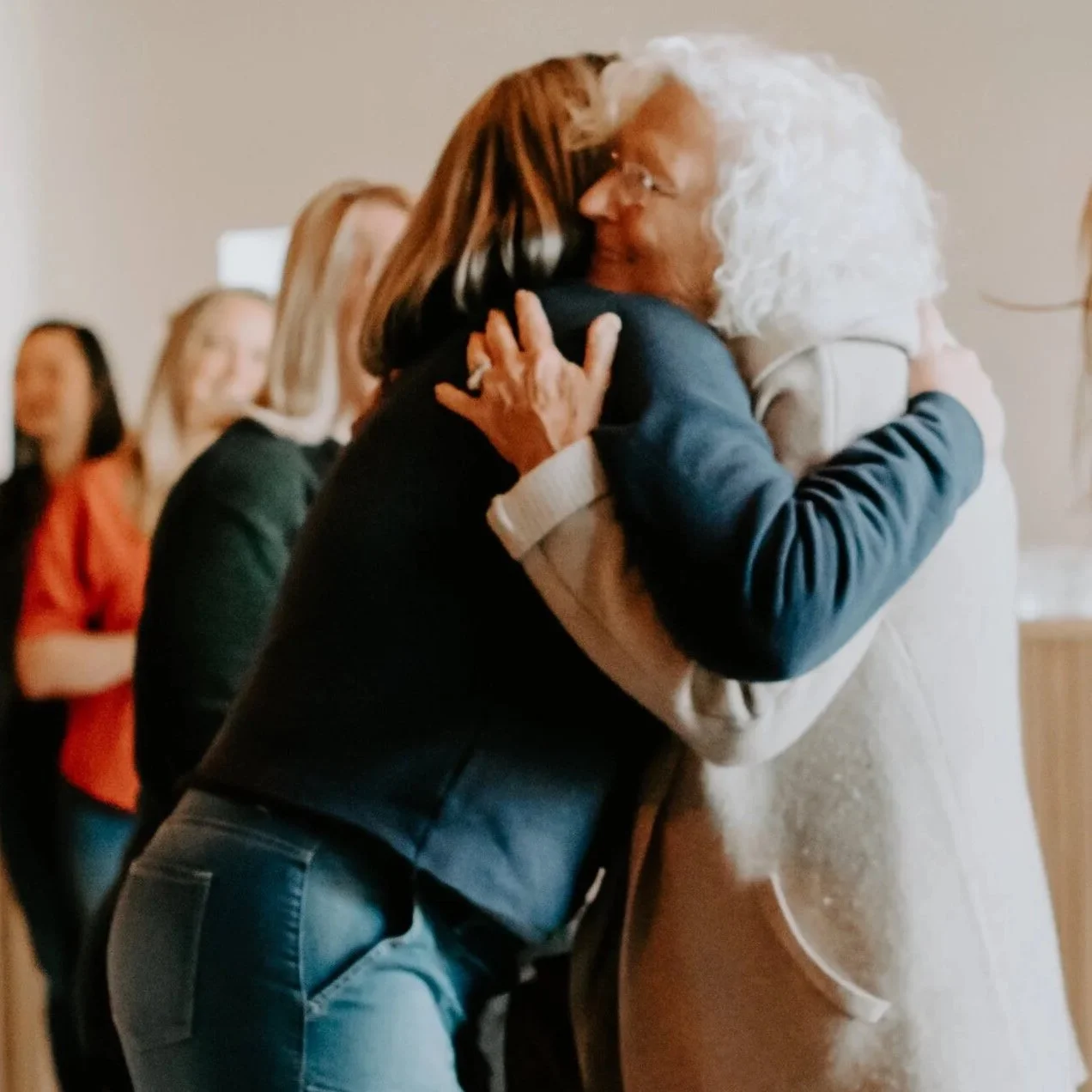Two women are hugging each other warmly in a friendly greeting, with one woman having gray curly hair and the other with straight brown hair, in an indoor setting with other people visible in the background.