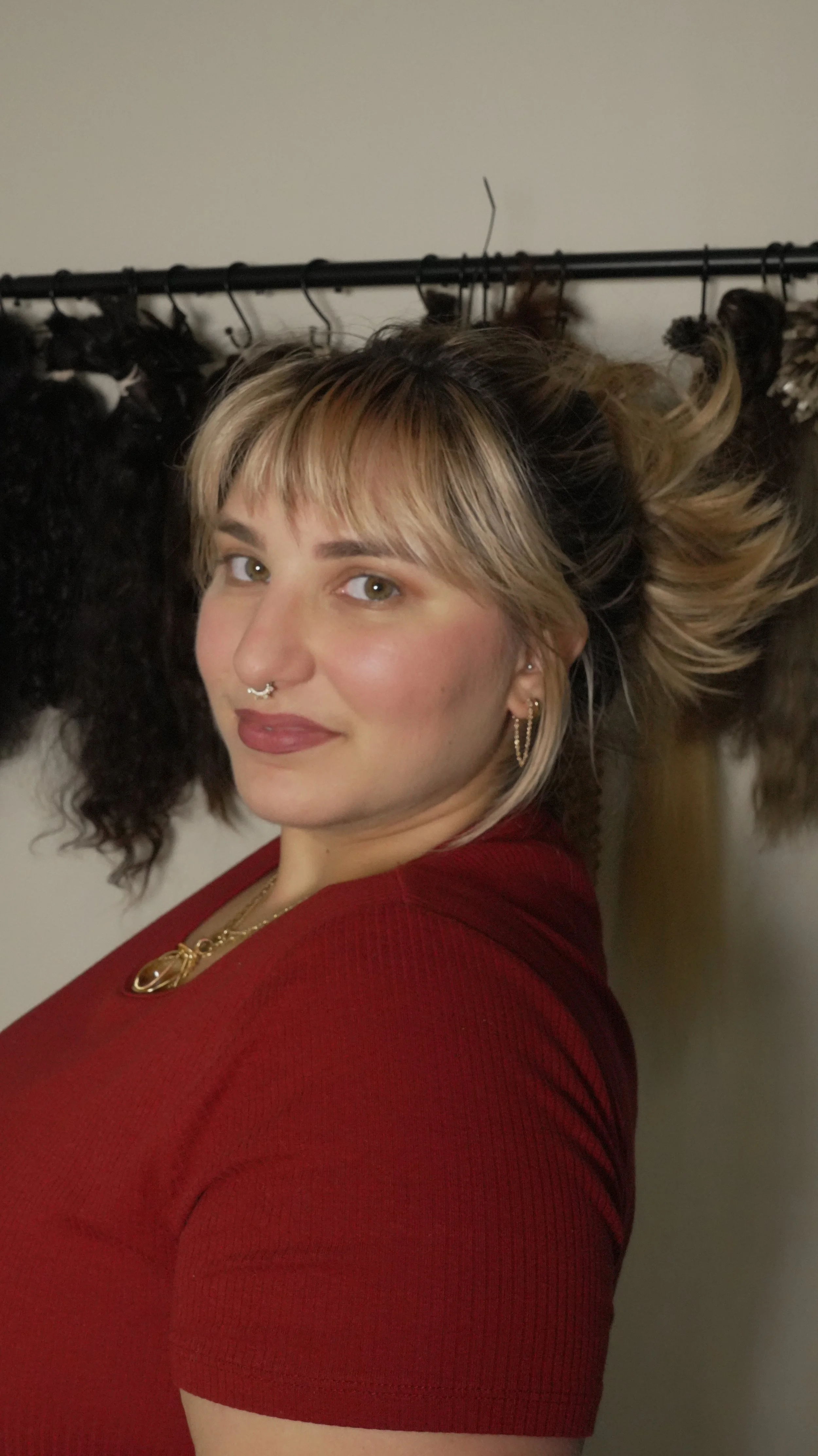 A woman with blonde hair, wearing a red top and gold jewelry, standing in front of a wall with raw unproccesed hair hanging on a rack.
