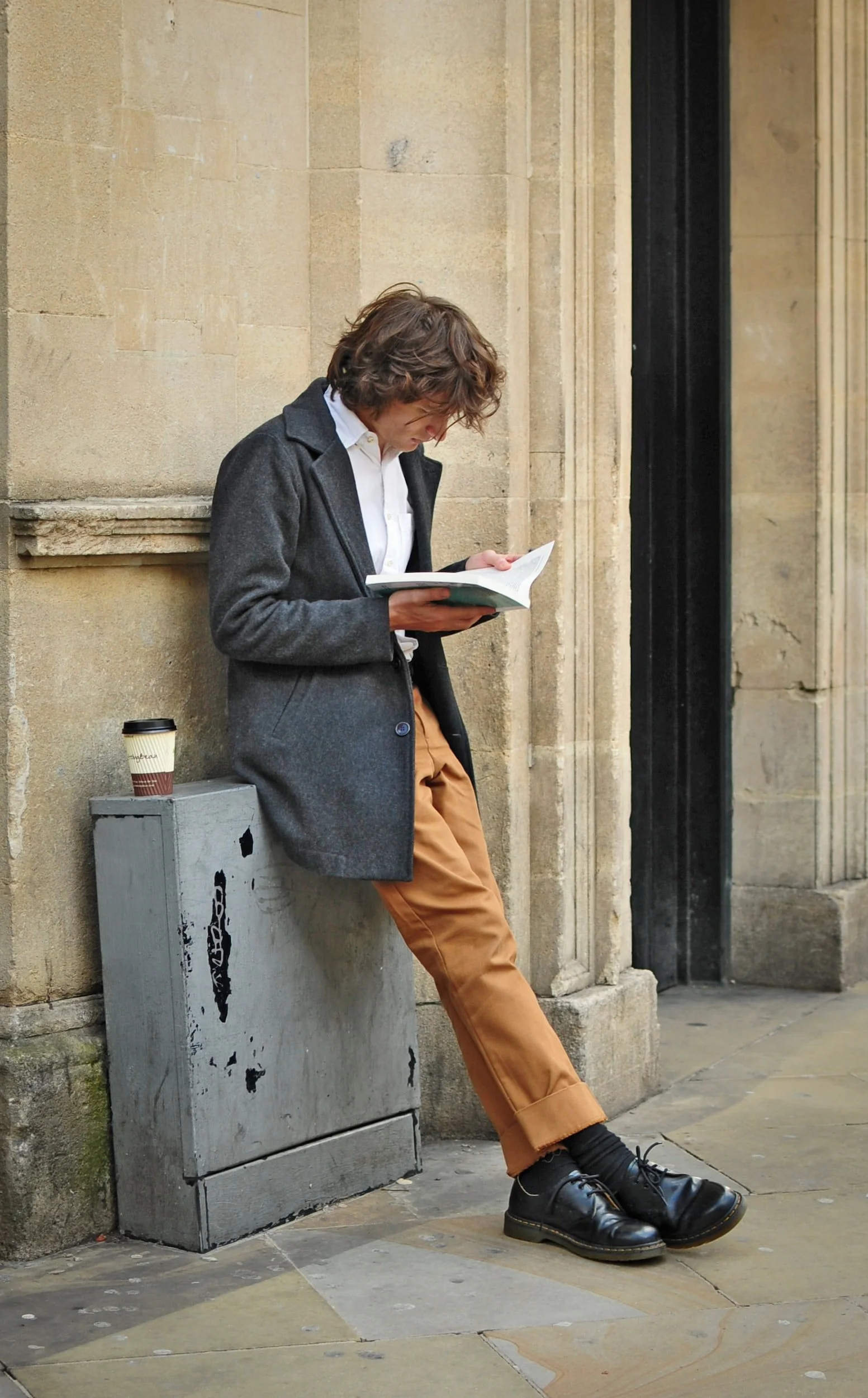 attractive Parisian man reading a book against an electrical box with a coffee sitting on it with old cream stone door frame he has long shaggy hair brown pants white button down and grey wool overcoat