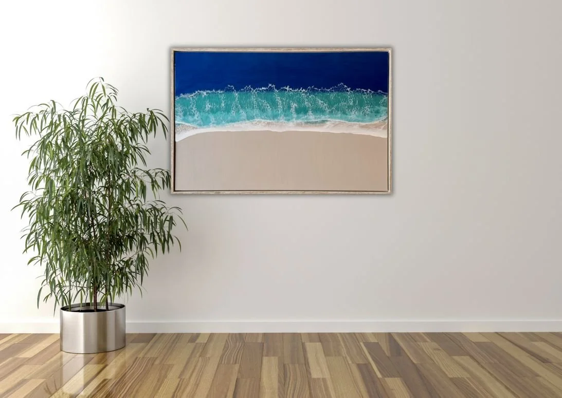 A framed painting of a beach scene with waves crashing onto the sand, hung on a white wall. To the left, there is a tall green plant in a metallic pot, and the floor is made of wood.