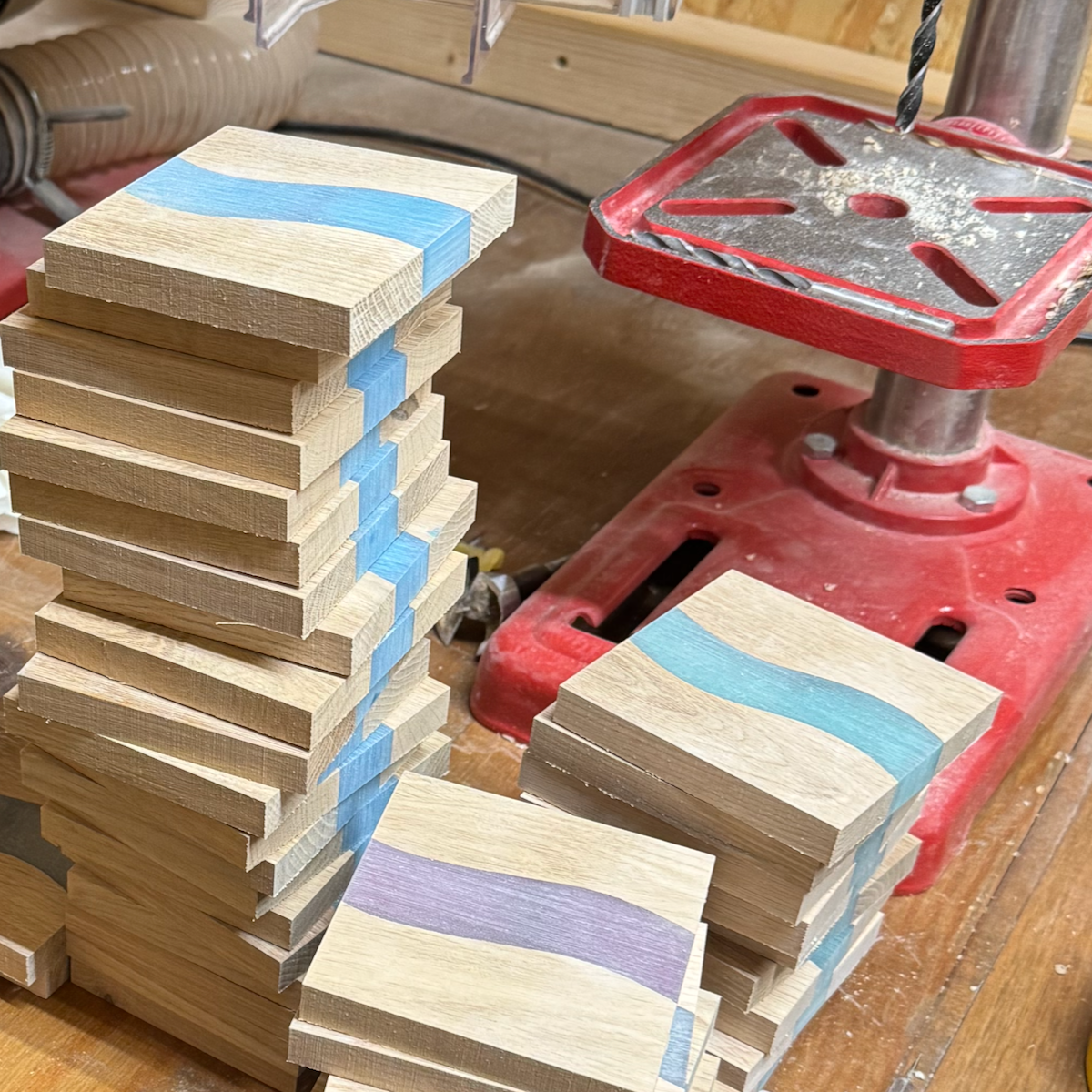 Stacked wood and resin coasters with blue, purple, and grey curved stripes, on a workbench with a red drill press nearby.