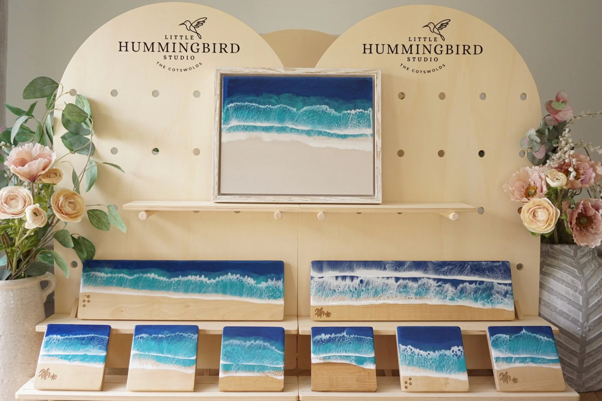 A display of ocean wave-themed resin artwork with floral arrangements on either side, at Little Hummingbird Studio in the Cotswolds.