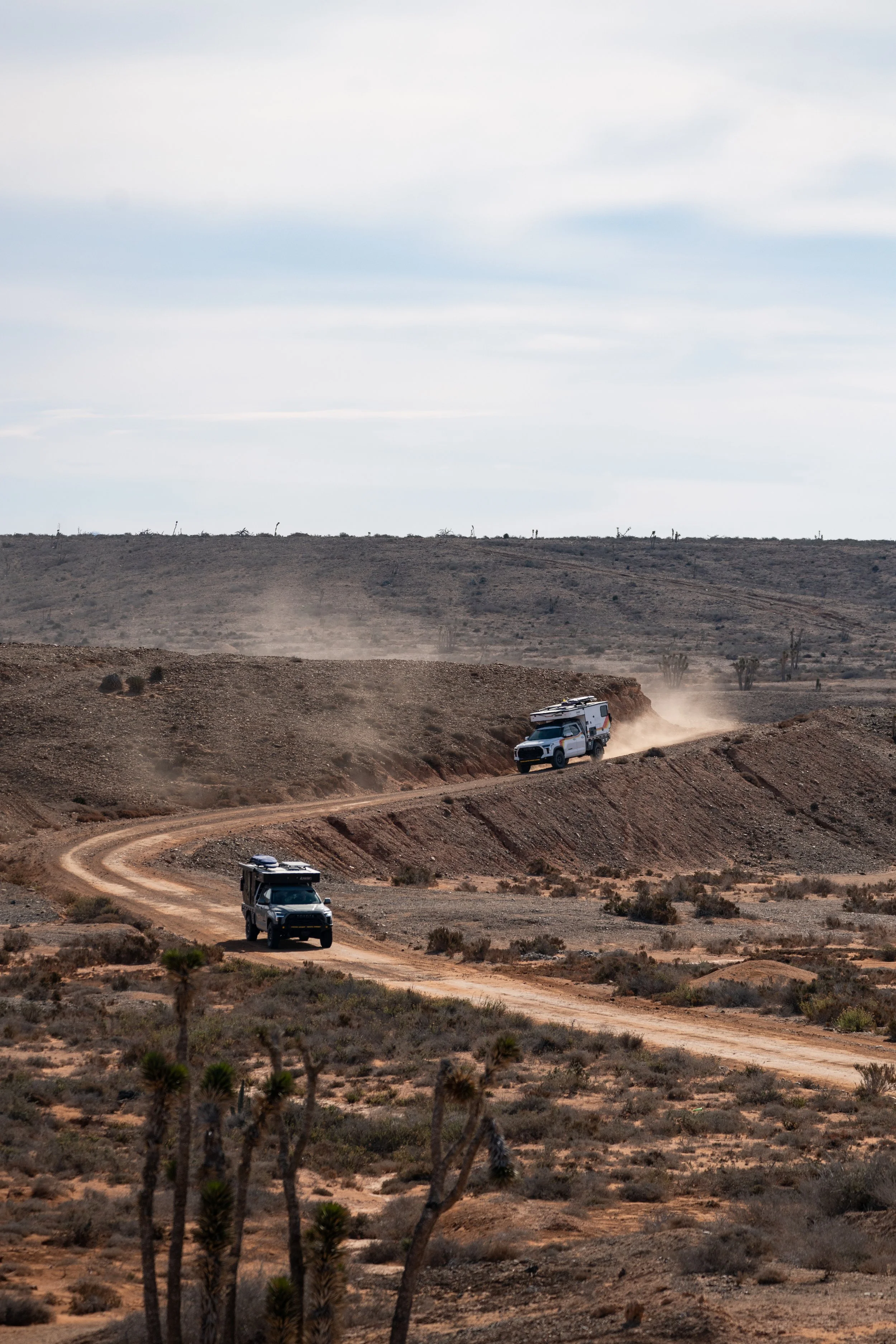 Two off-road vehicles driving on a winding dirt road in a desert landscape with sparse vegetation and a hill in the background under a cloudy sky.