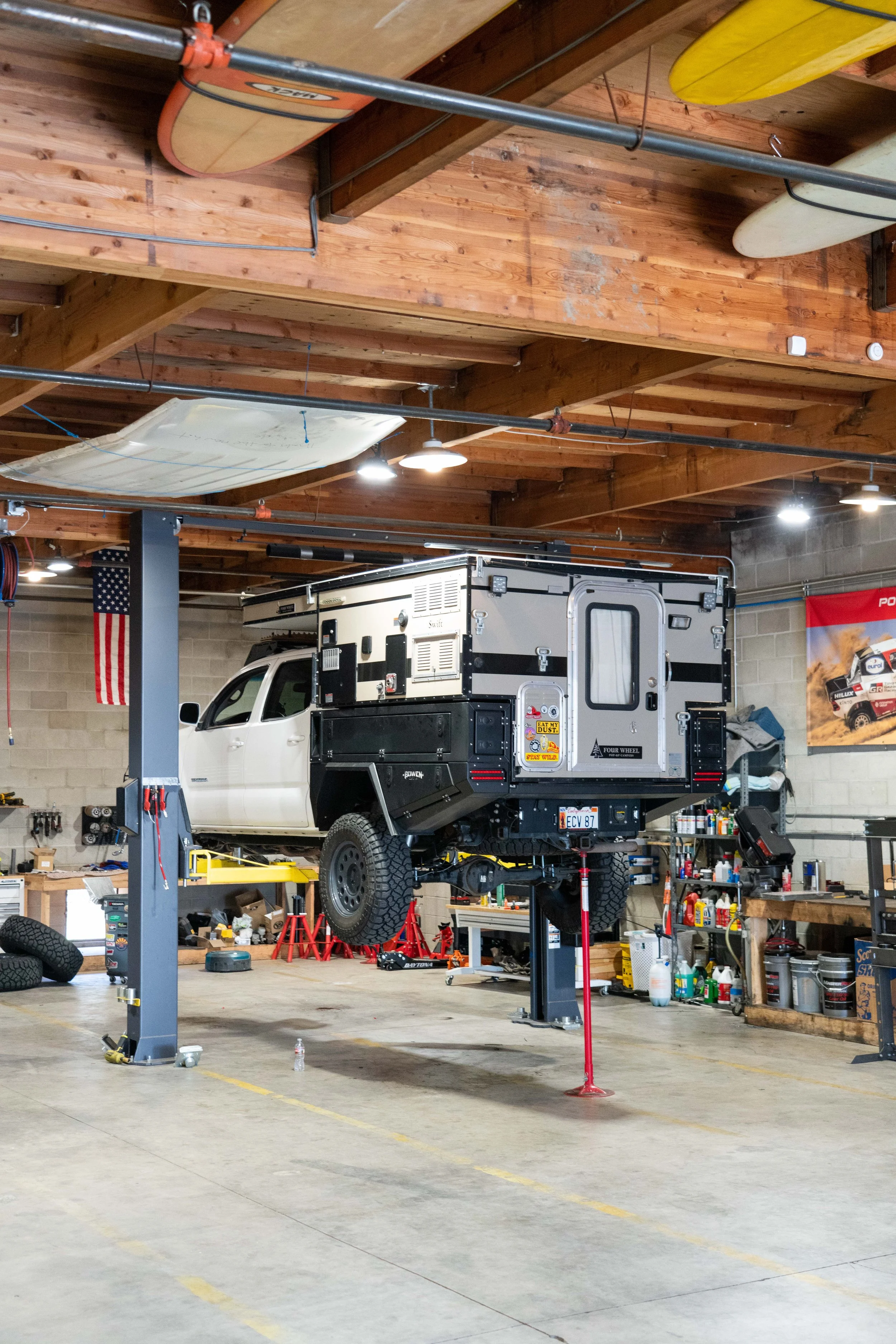 A truck is lifted on a hydraulic lift inside a garage workshop, with various tools, shelves, and surfboards stored on the ceiling and walls.