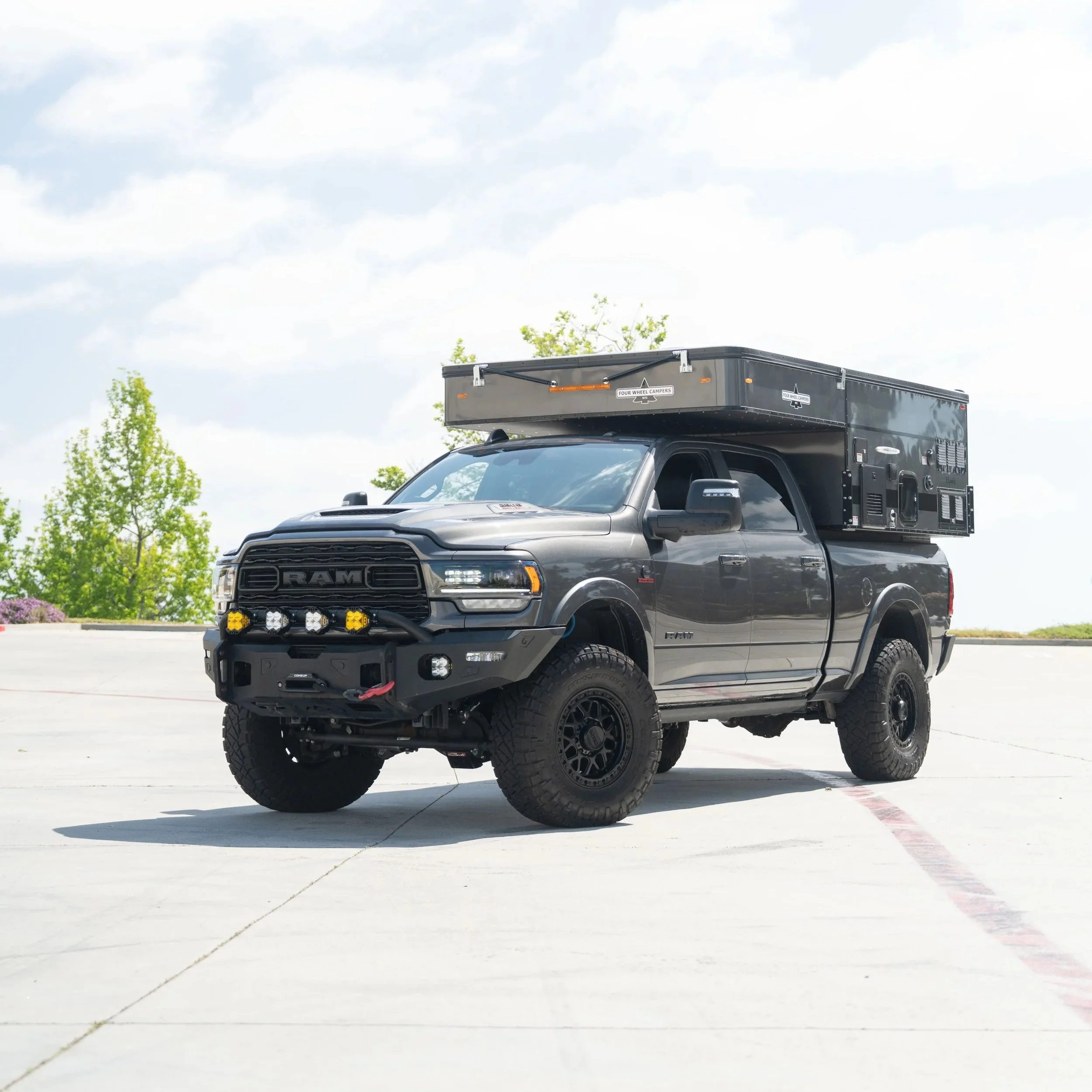 Black pickup truck with off-road tires and a black camper shell on top, parked on a concrete surface under a partly cloudy sky, with trees in the background.