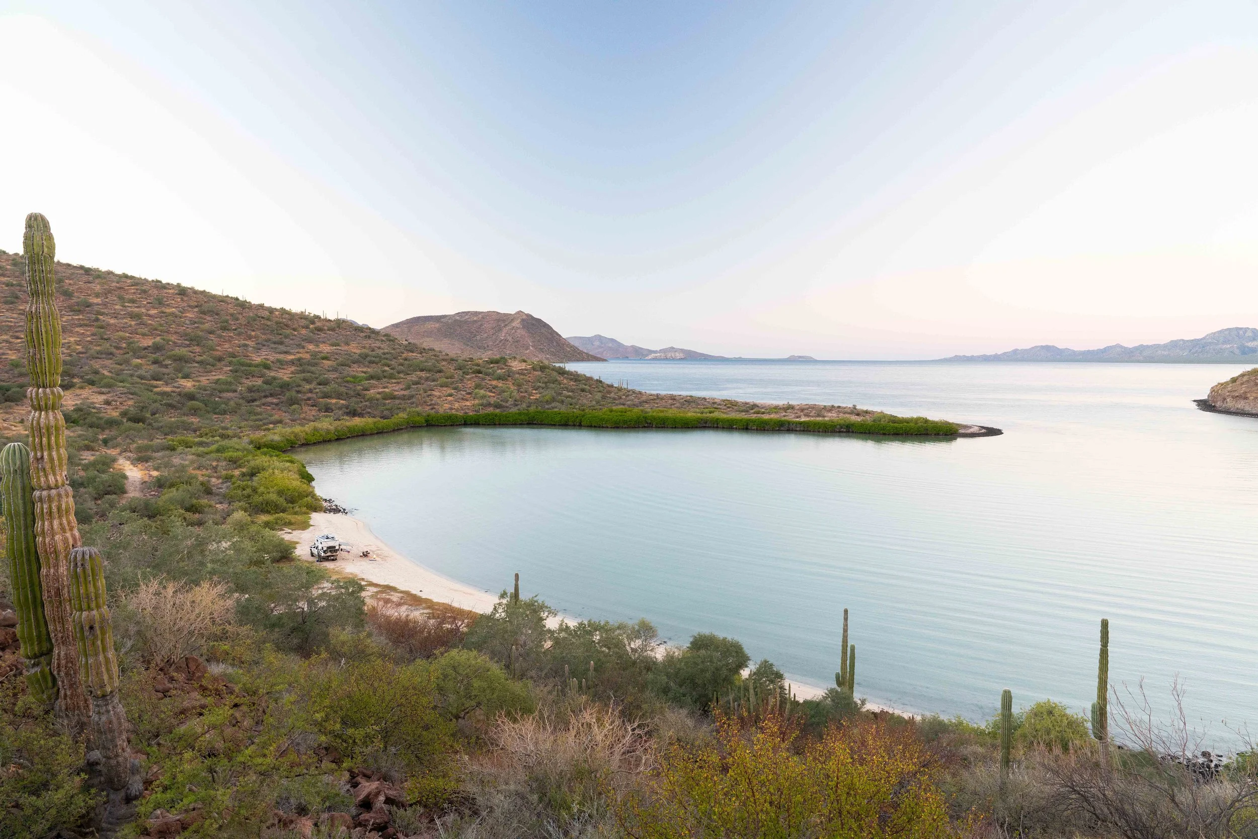 A desert landscape with cacti and sparse bushes, a small beach with a parked vehicle, and a large body of water with distant mountains in the background.