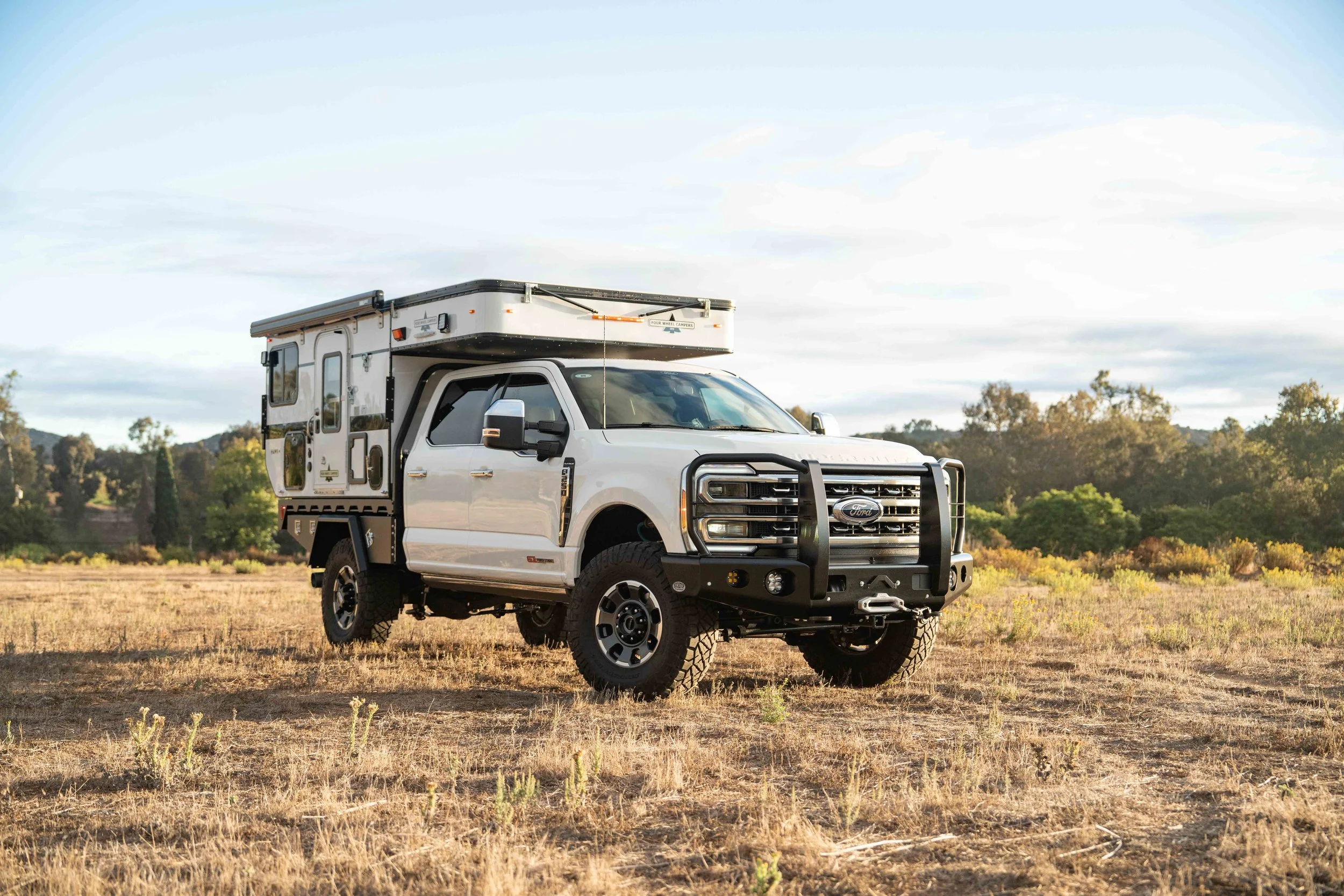 White pickup truck with a camper shell and rooftop tent parked on a grassy field with trees and hills in the background.
