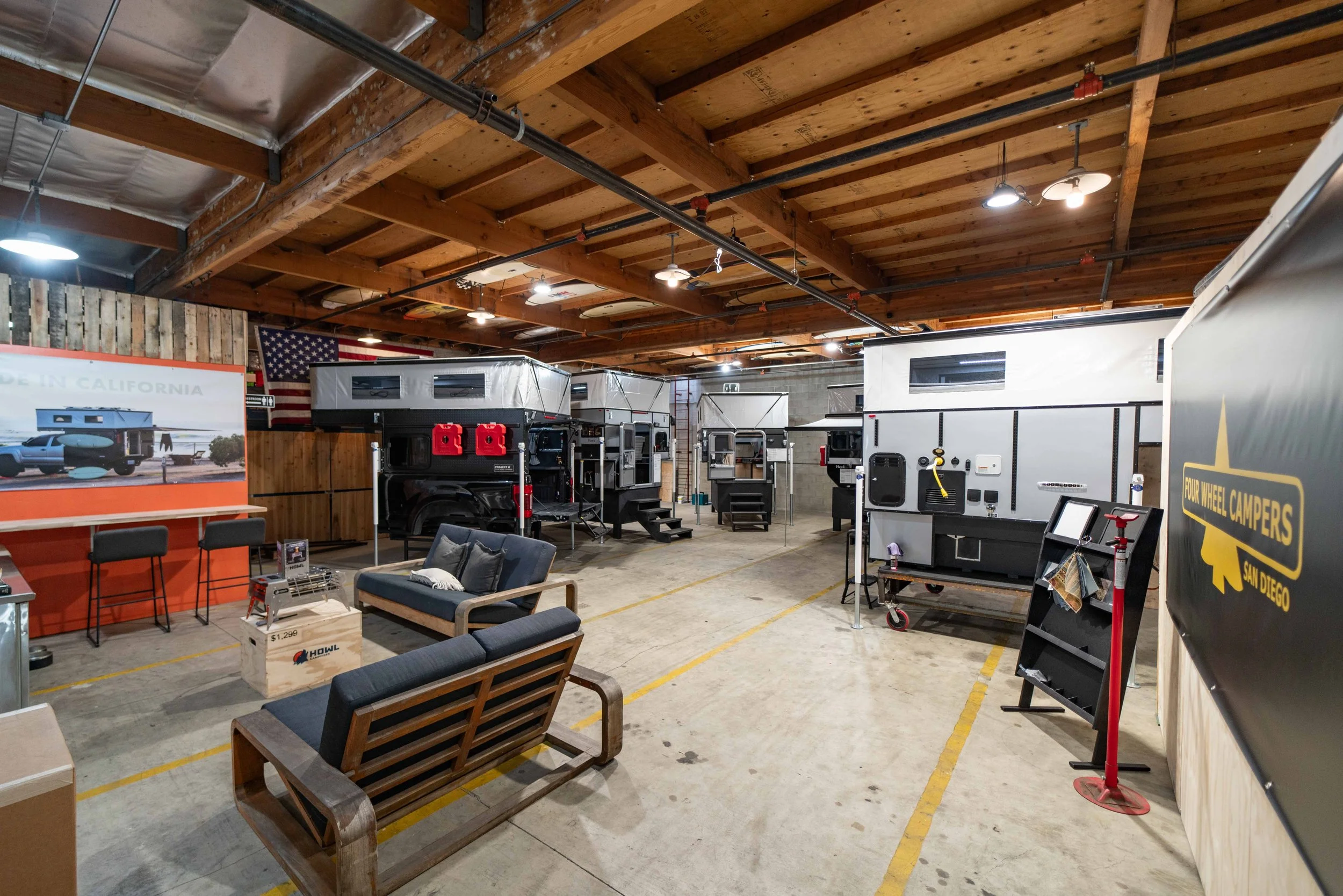 Indoor showroom or store displaying various camper trailers and RVs with wooden ceiling, American flag, and seating area.