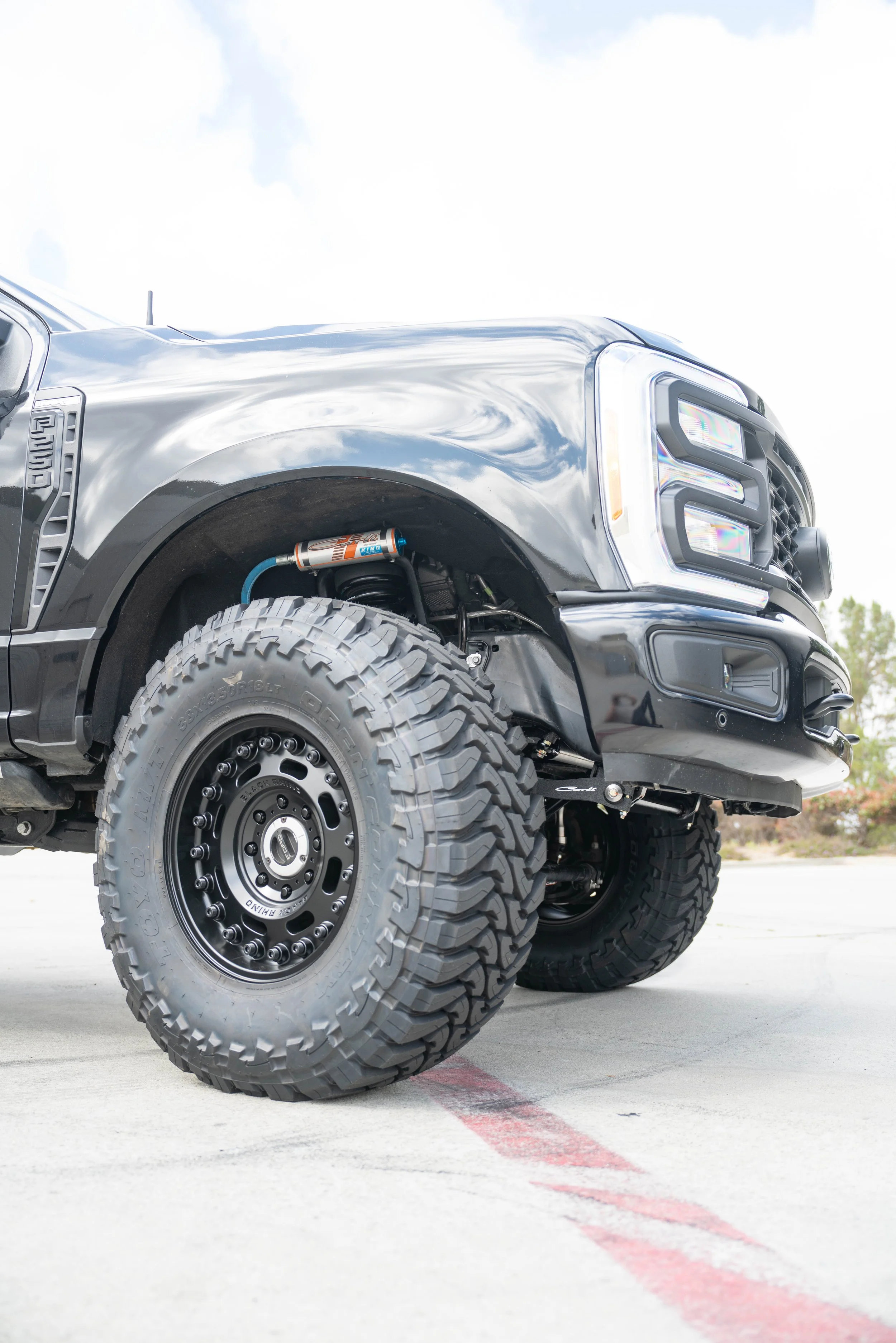 Close-up of the front part of a black off-road pickup truck, with large tires, a suspension lift, and a visible shock absorber, parked on a concrete surface.