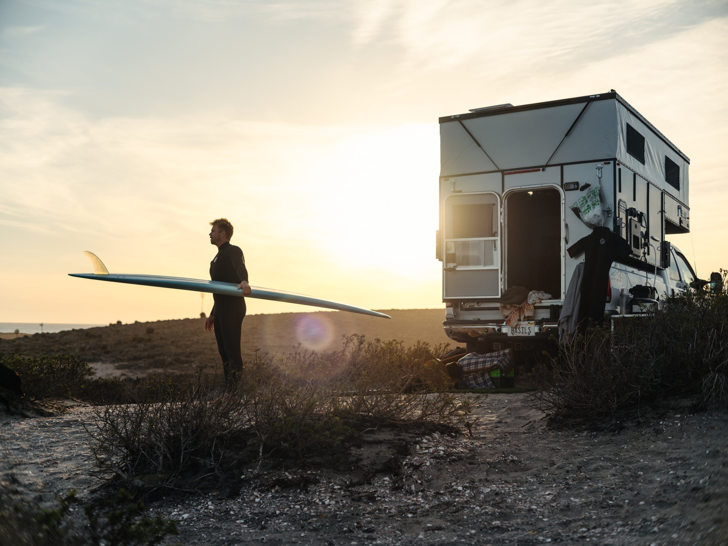 A man is walking on a beach holding a surfboard, with a camper van parked nearby during sunset or sunrise.