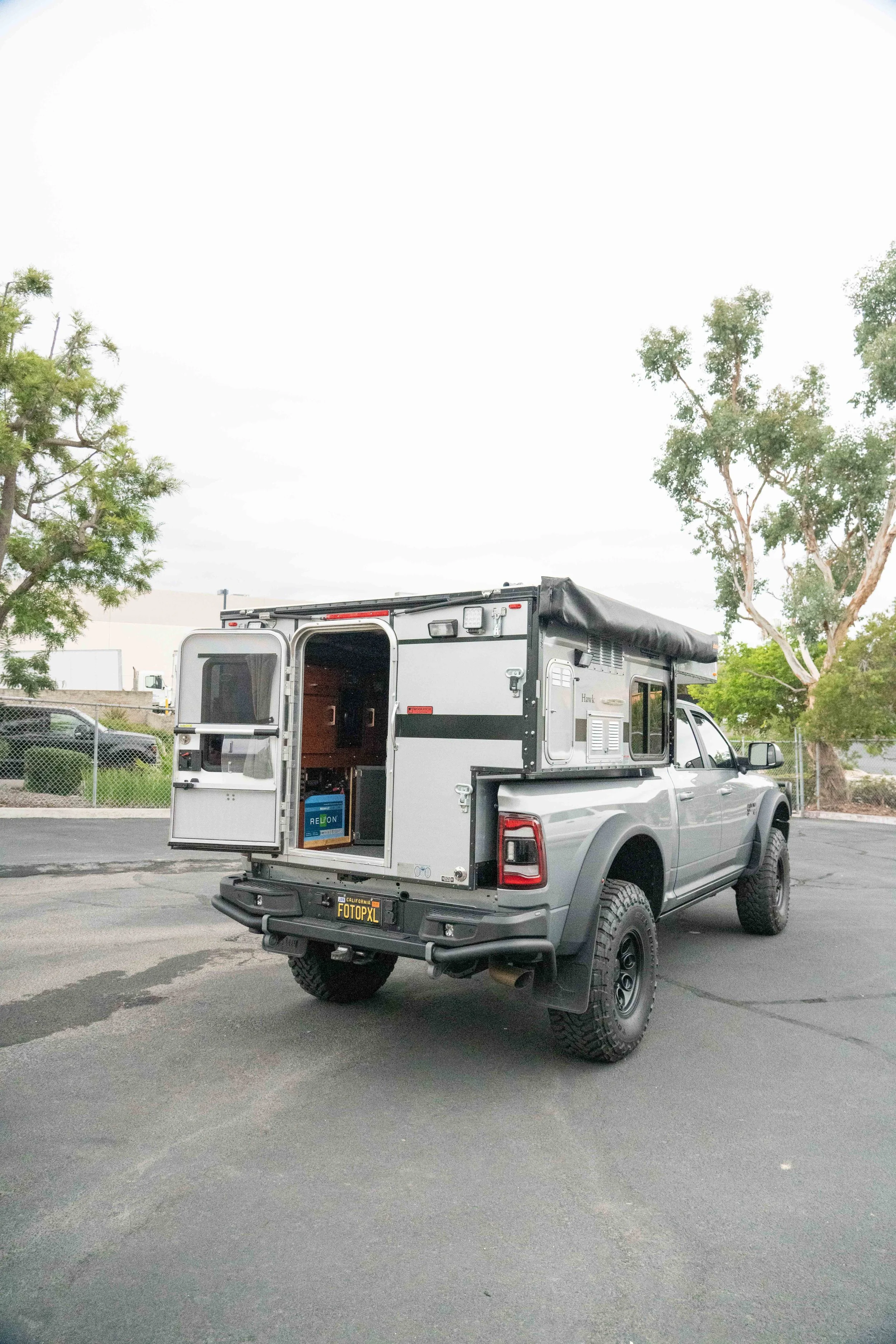 A silver pickup truck with a camper trailer attached, parked in an outdoor lot with trees and a chain-link fence in the background.