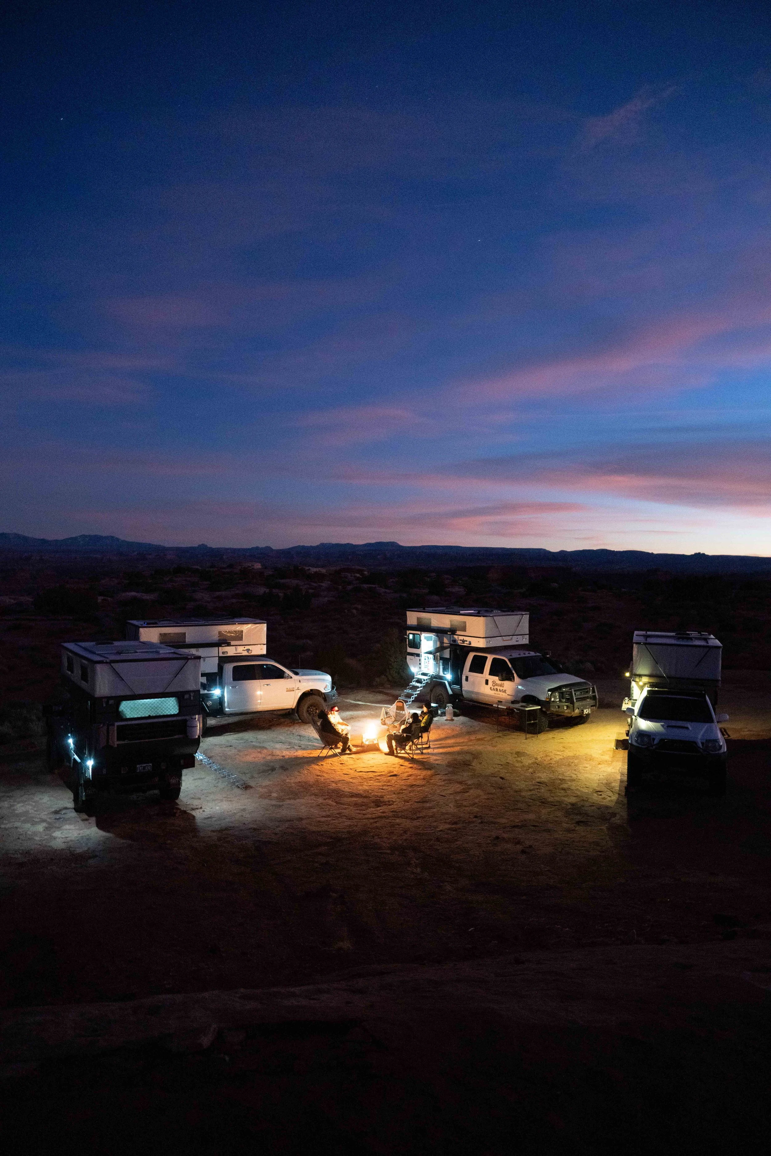 An outdoor camping scene at dusk with several RV trailers and a group of people sitting around a campfire. The sky shows a gradient of colors from purple to pink and blue, with some clouds and stars visible.