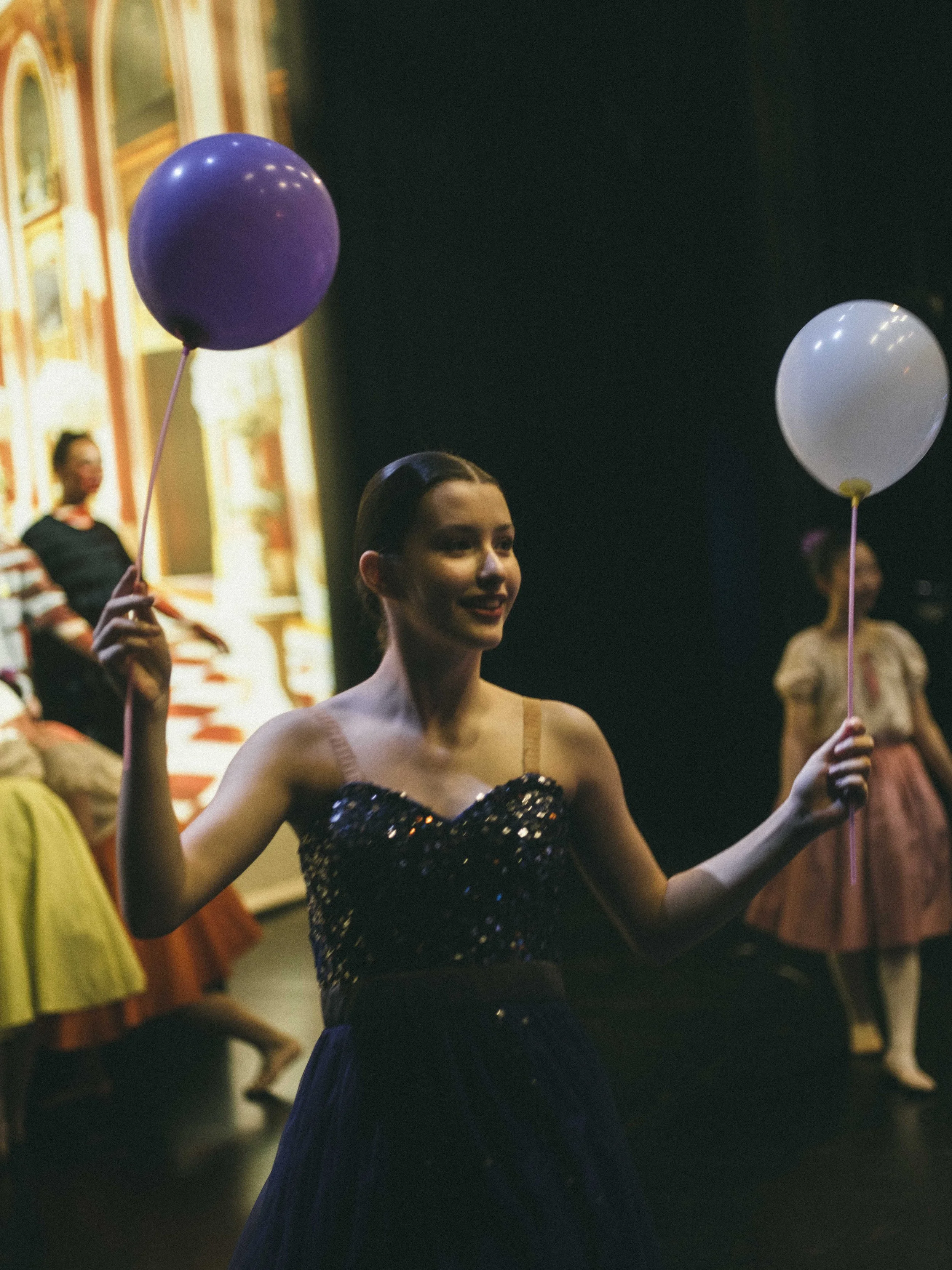 A woman holding a purple balloon in her right hand and a white balloon in her left hand, standing on a stage with other women in vintage dresses in the background.