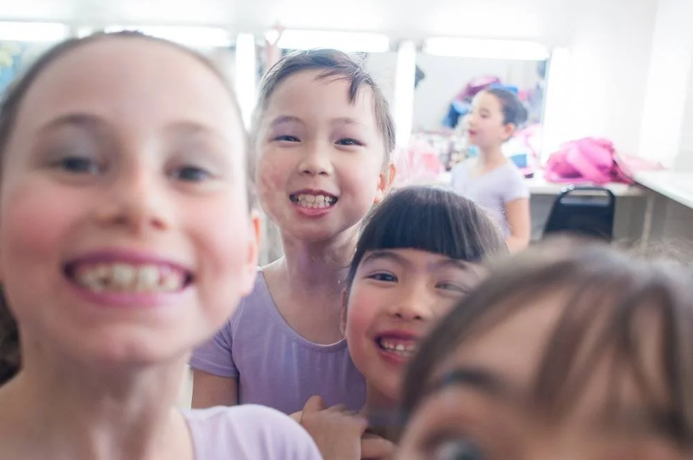 Four children smiling and posing for a close-up photo indoors, with a fifth child in the background near a table.