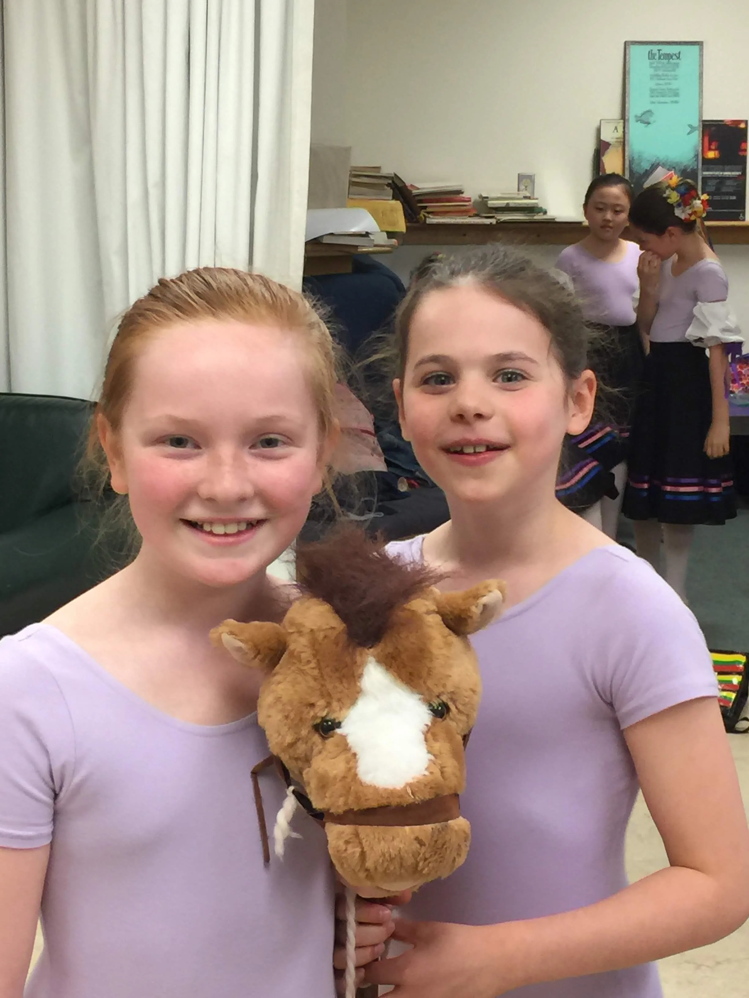 Two young girls in lavender costumes smiling and holding a plush horse puppet with brown fur, white forehead marking, and a mane, in a room with others in costumes in the background.