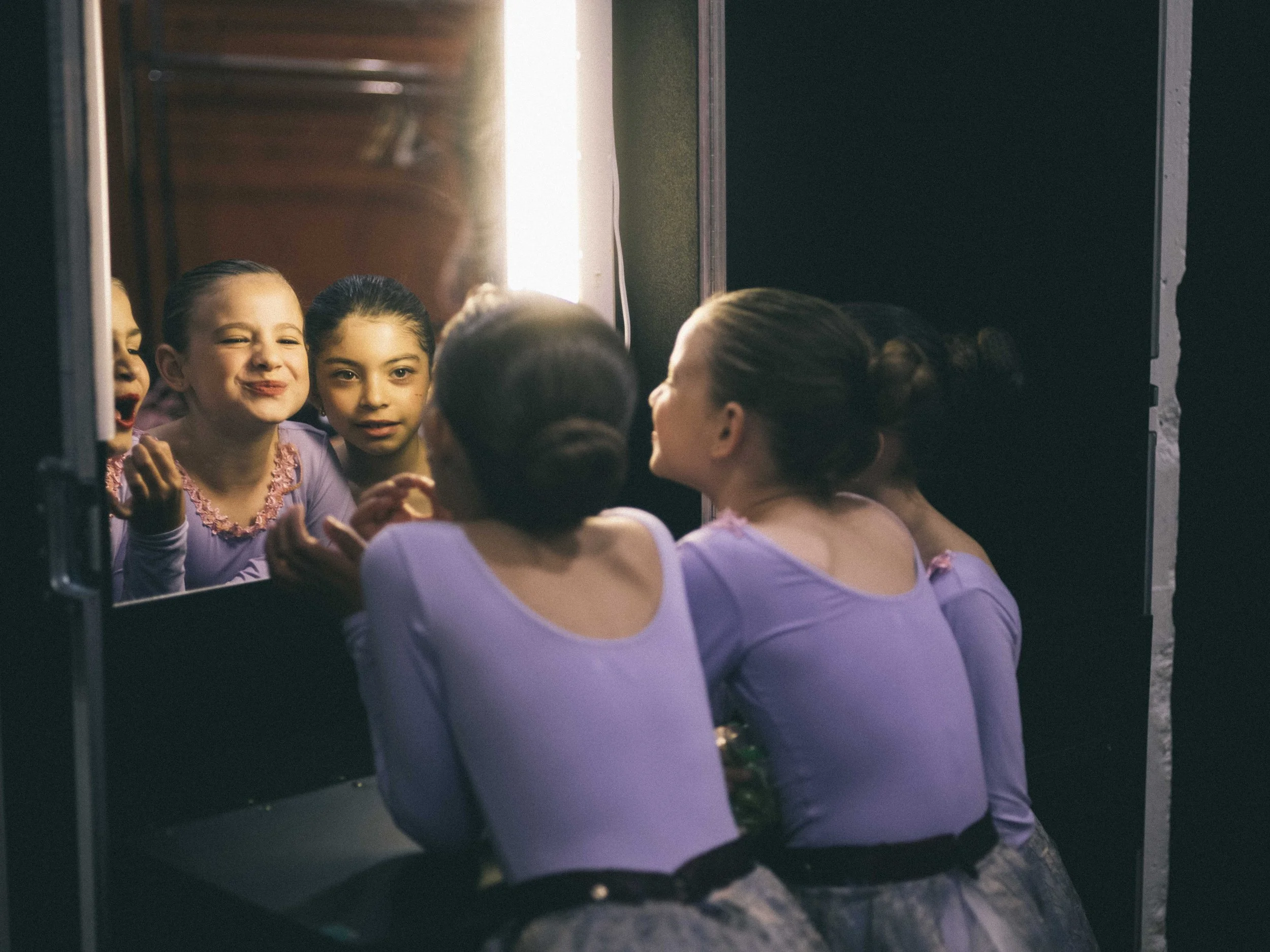 A group of young ballerinas in lavender costumes looking at themselves in a mirror, with one girl making a silly face.