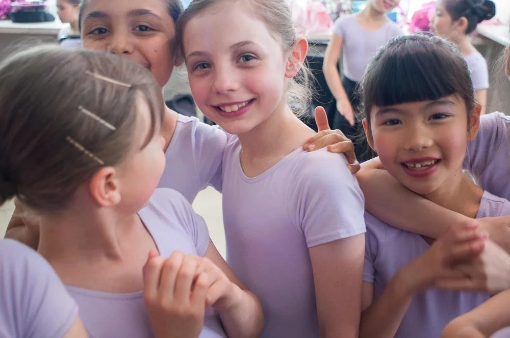 Group of four young girls smiling and hugging each other, wearing light purple shirts, outdoors, with other children in the background.