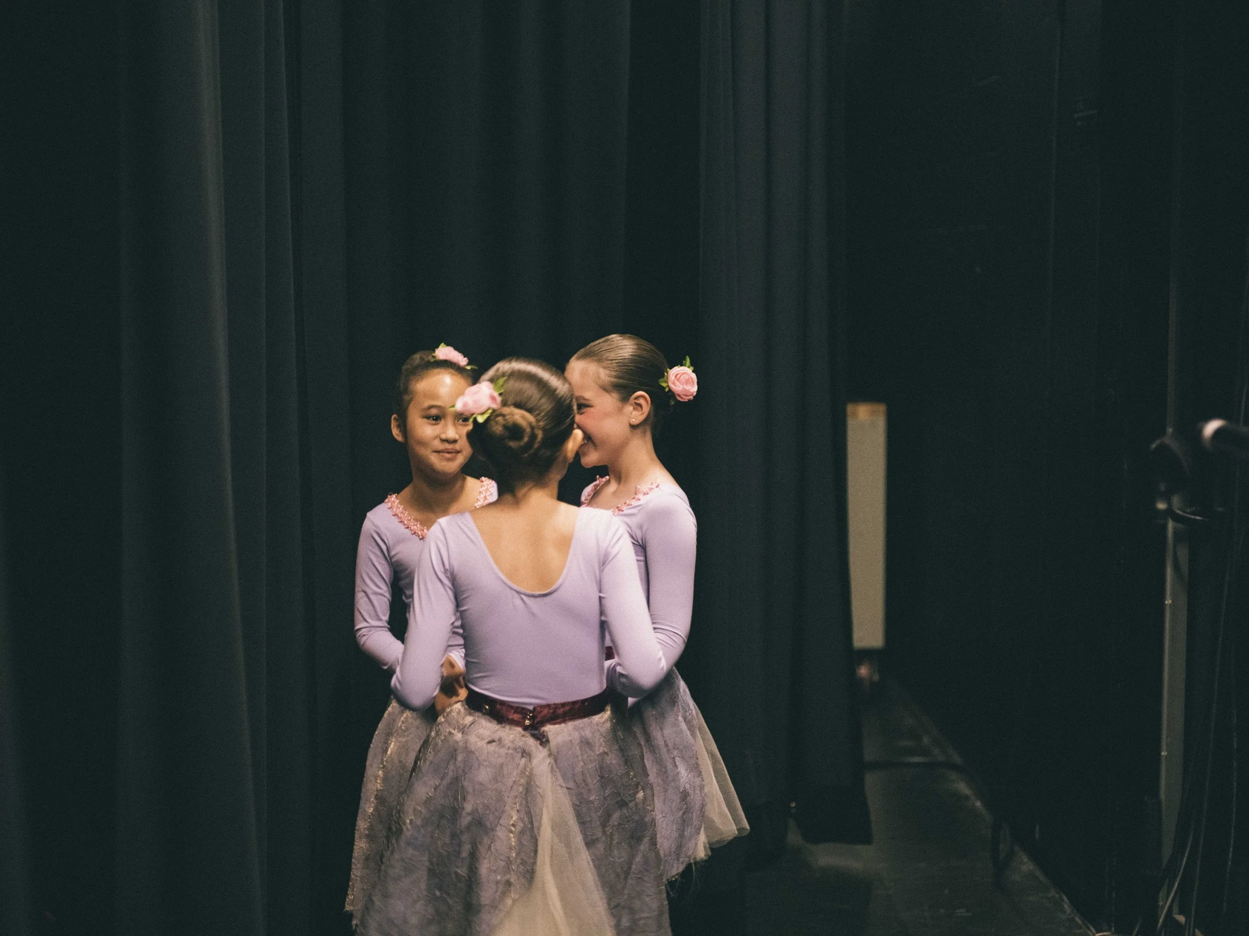 Three young ballet dancers in lavender costumes with floral headpieces backstage.