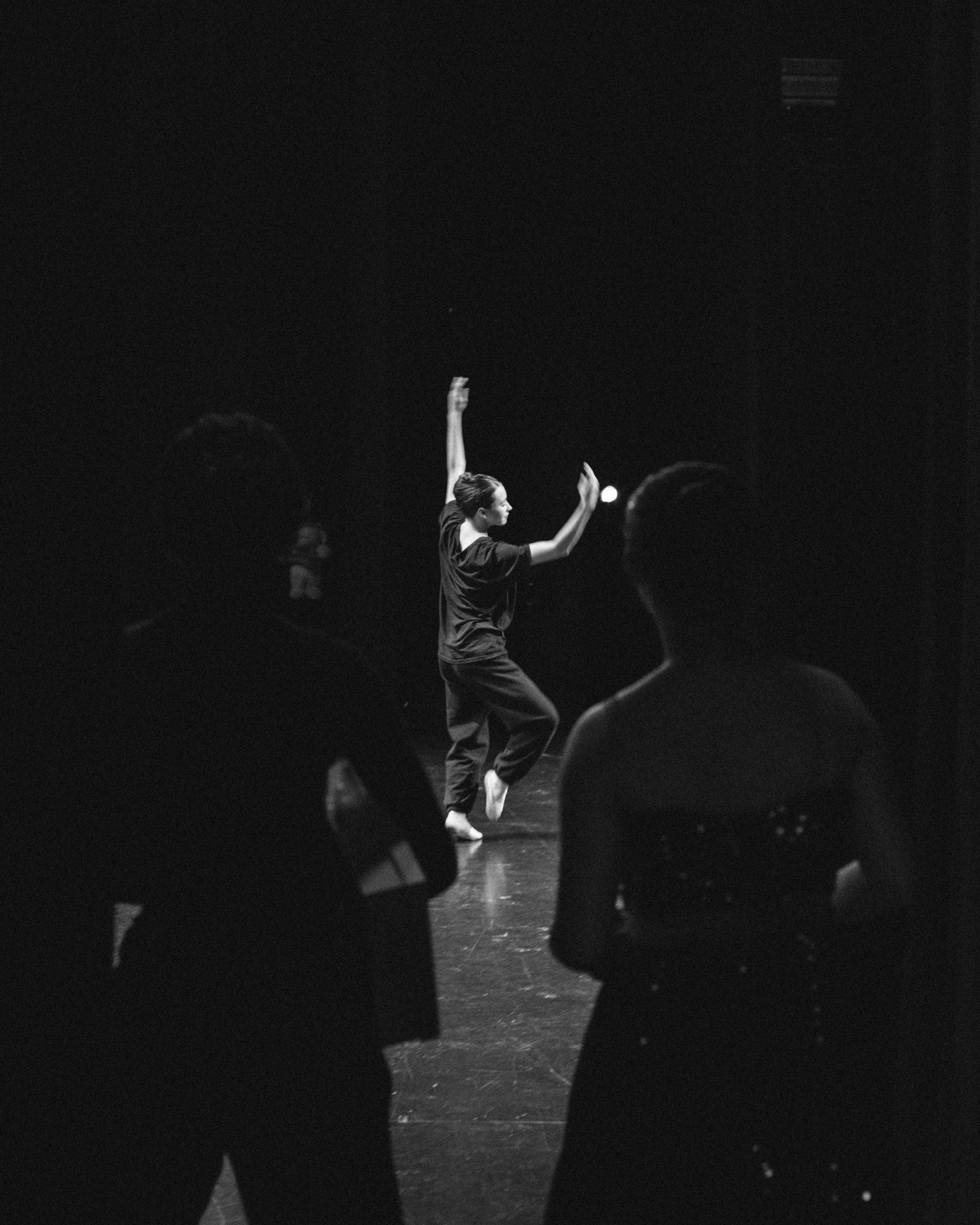 A woman dancing on a stage, seen from behind and to the side, with two people watching in the foreground.
