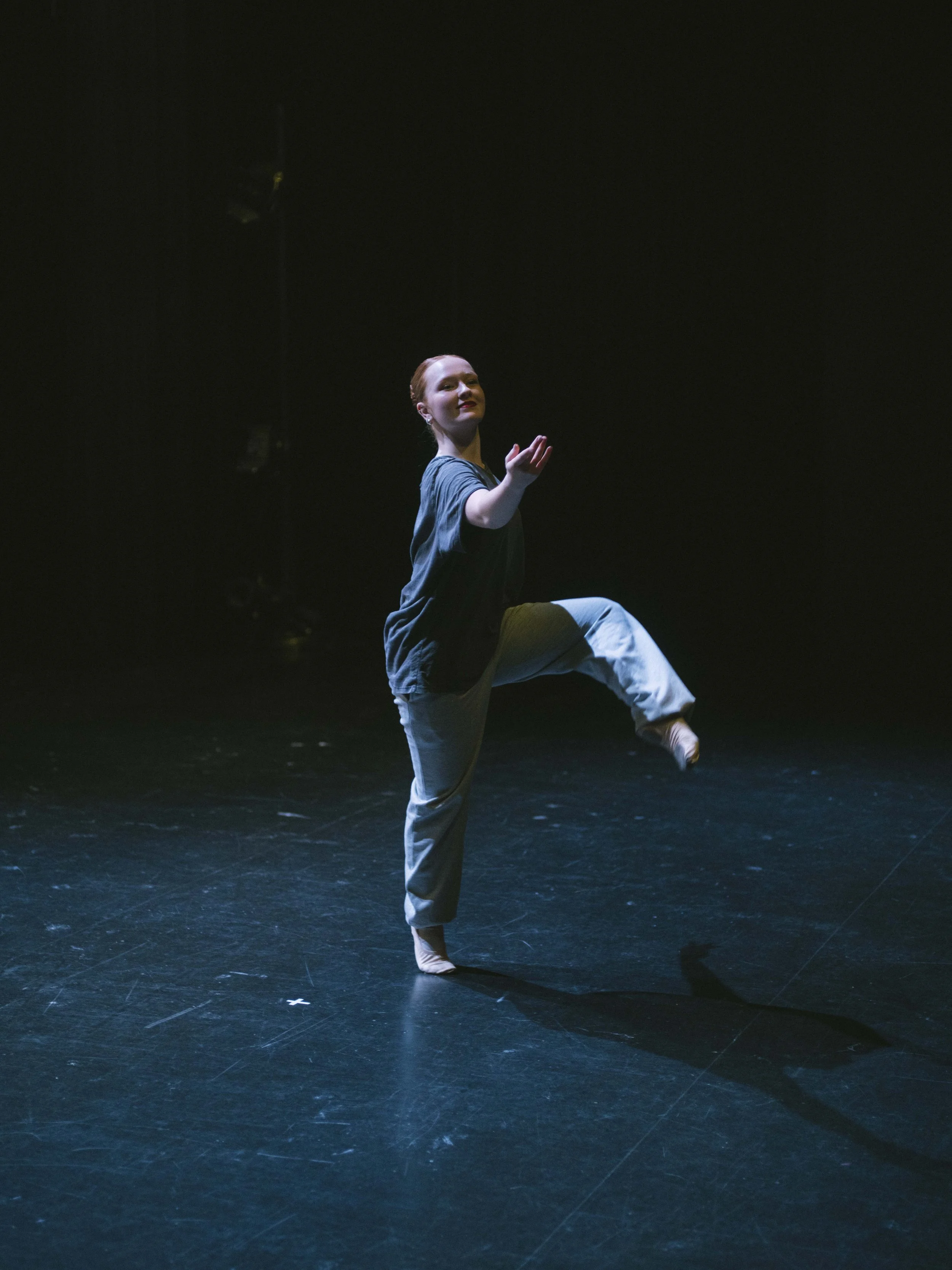 A young woman performing a dance move on a dark stage, standing on one leg with her other leg bent and raised, and raising one arm with an open hand.