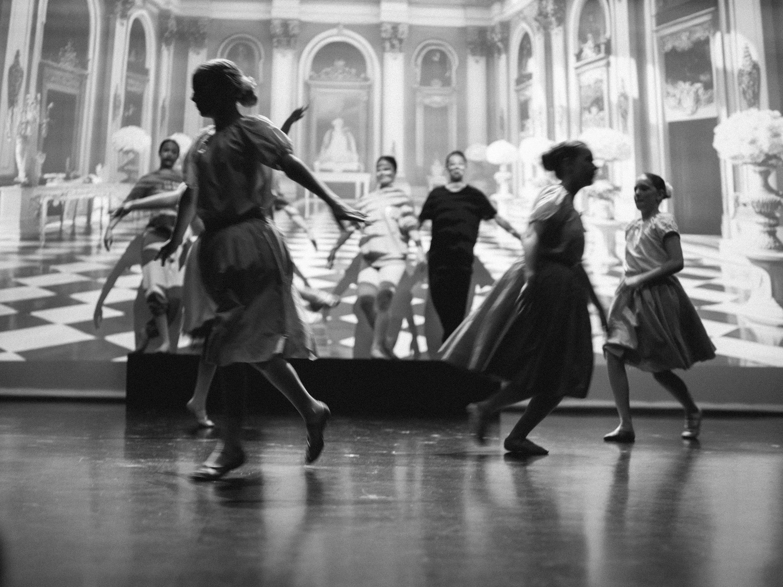 Black and white photo of a theatrical dance performance with multiple dancers in vintage costumes on stage, with a detailed palace-like backdrop.