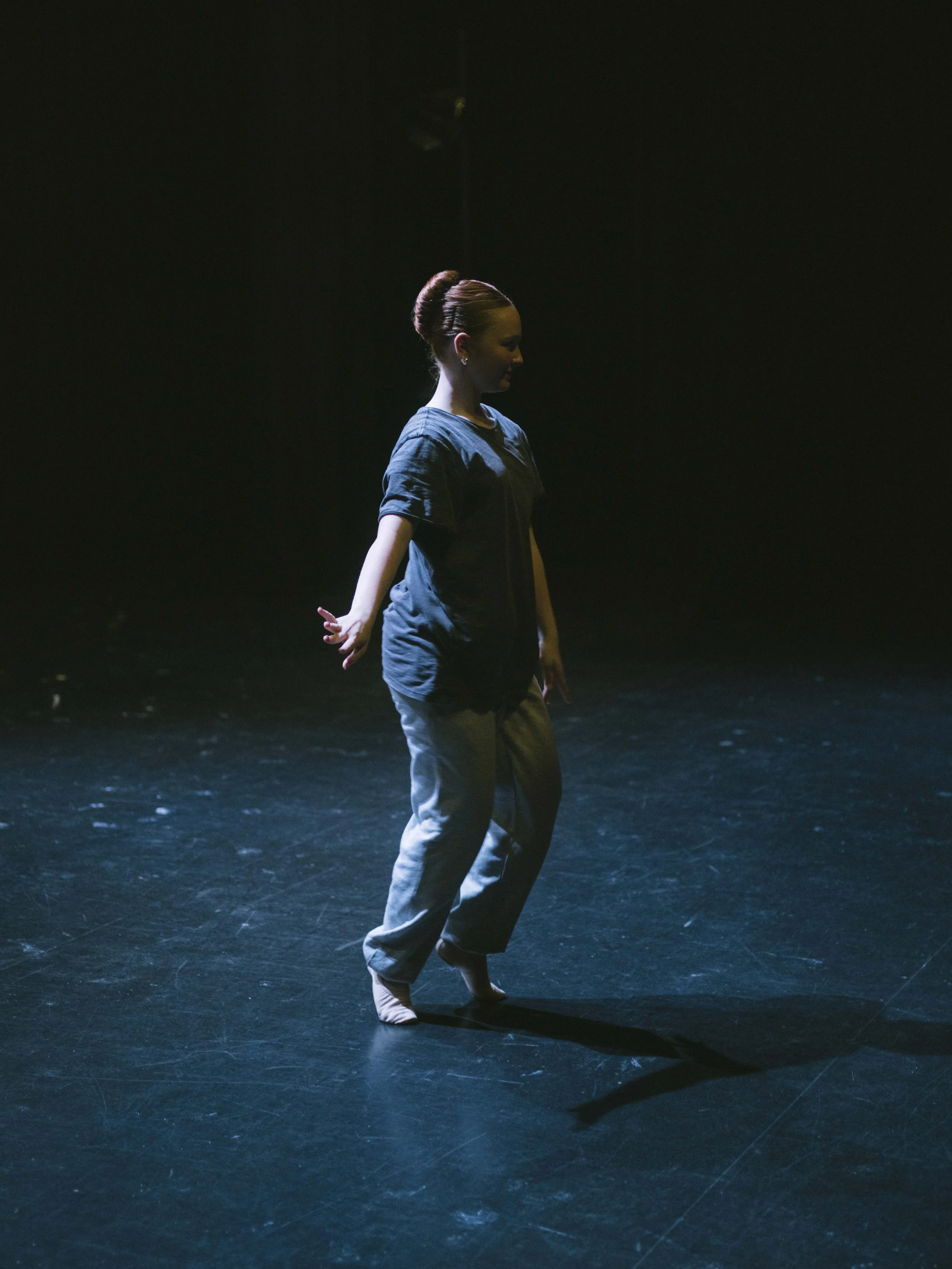 A woman standing on a stage with her eyes closed, wearing a dark blue top and beige pants, with her hair styled in an elegant bun, under a spotlight.