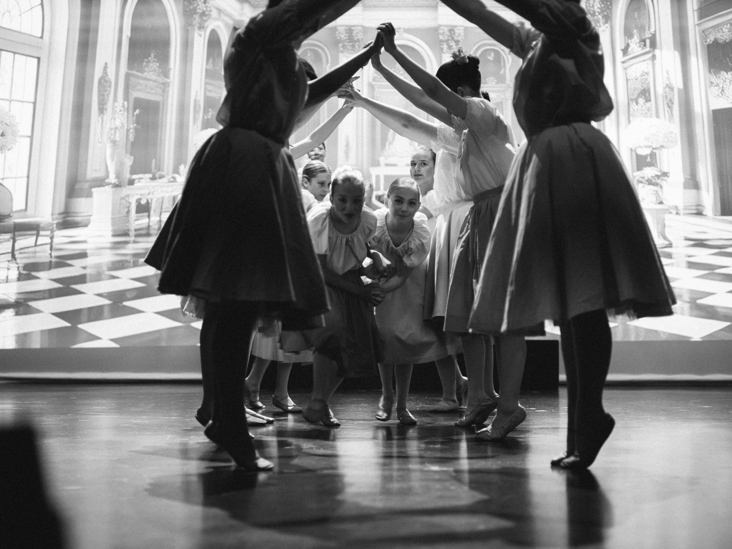 A group of young girls performing a dance on stage, forming a circle with some standing and others crouching, with a large screen or backdrop behind them depicting an ornate room with patterned flooring.