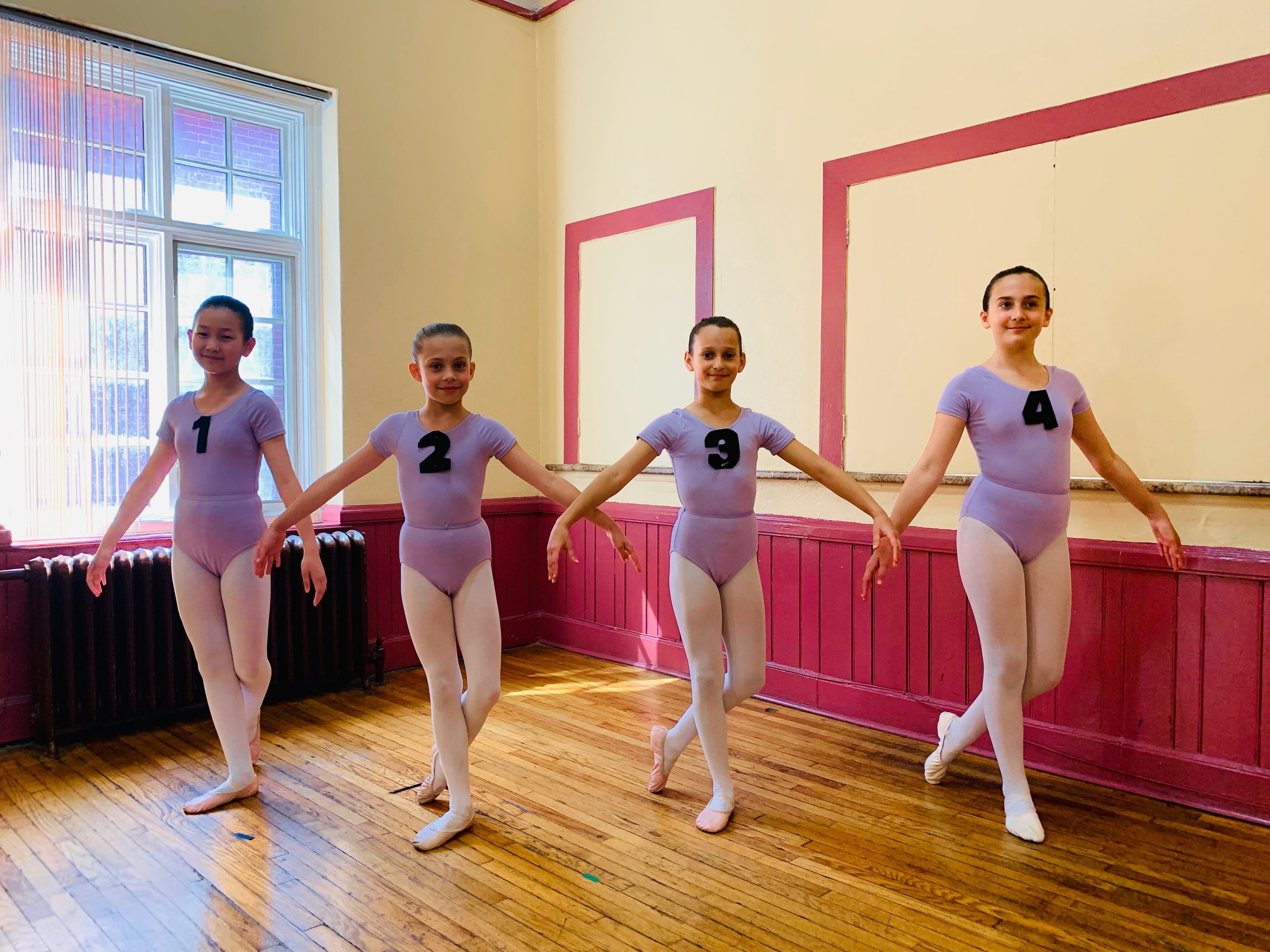 Four young ballet dancers standing in a line with their arms extended, wearing lavender leotards, tights, and ballet slippers, with numbered black symbols from 1 to 4 on their chests, inside a dance studio with hardwood floors and a large window.