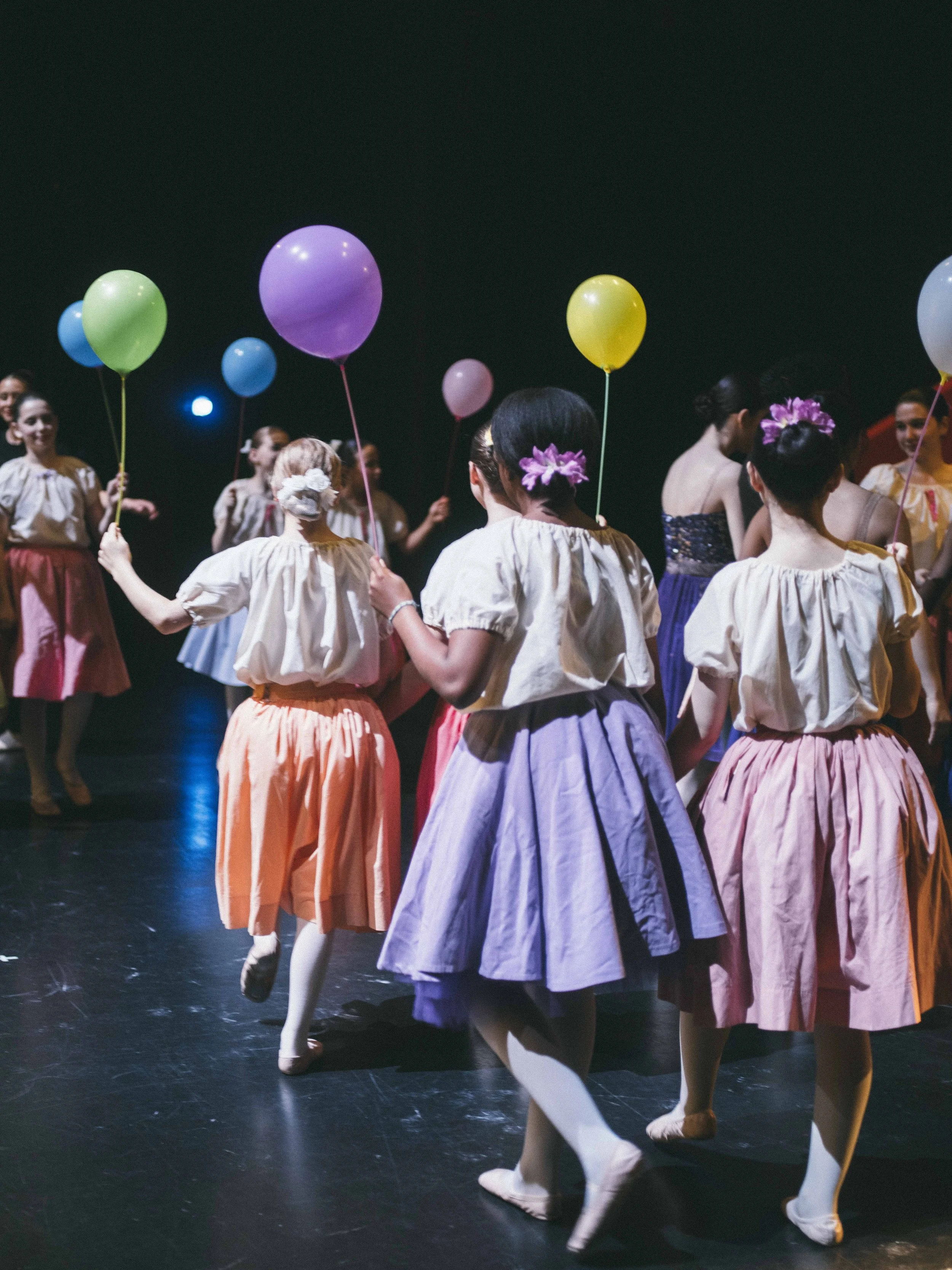 Girls in colorful skirts and blouses holding balloons, dancing on a stage during a performance.