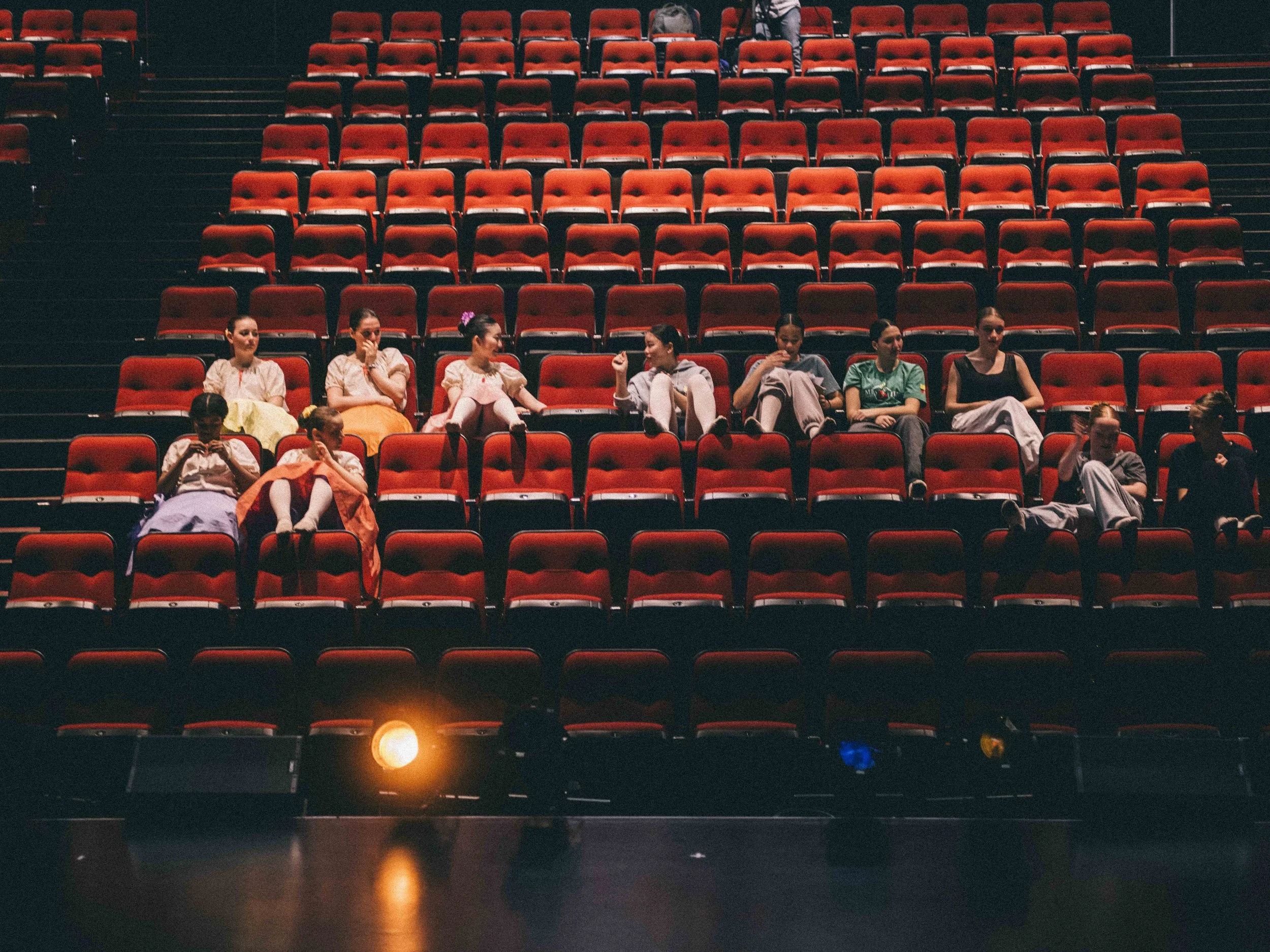 Group of children seated in red theater seats, some engaged in conversation, in an auditorium or theater setting.