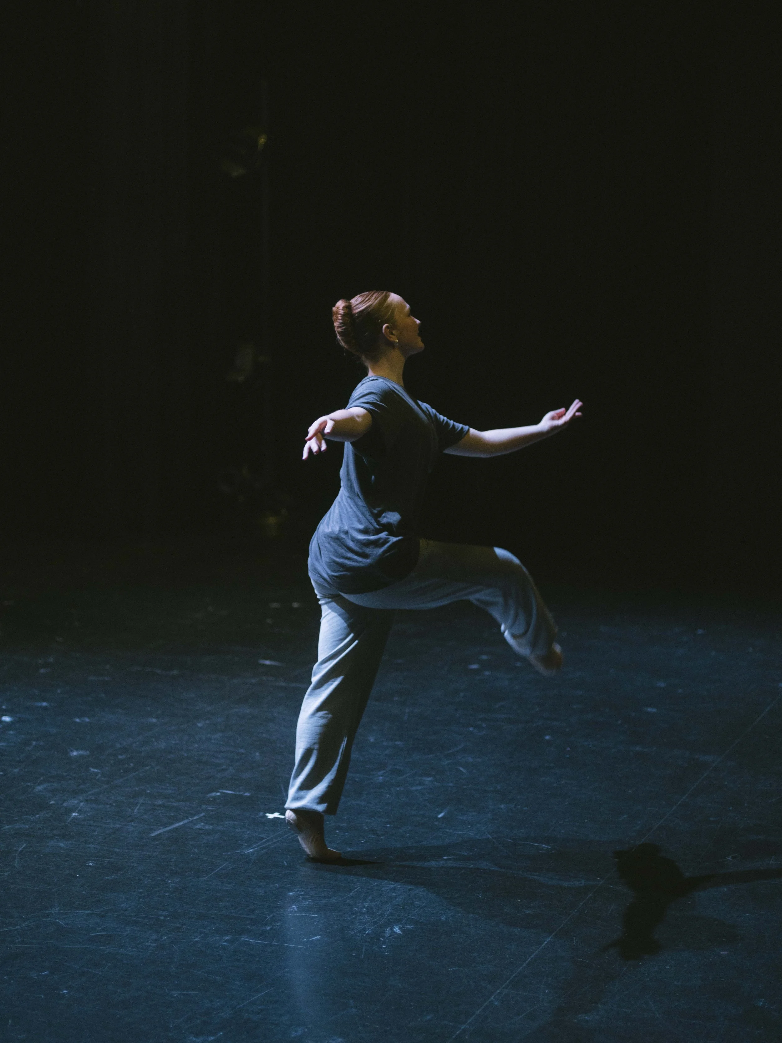 A woman performing a dance move on stage, standing on one leg with arms extended, in a dark setting with spotlight lighting.