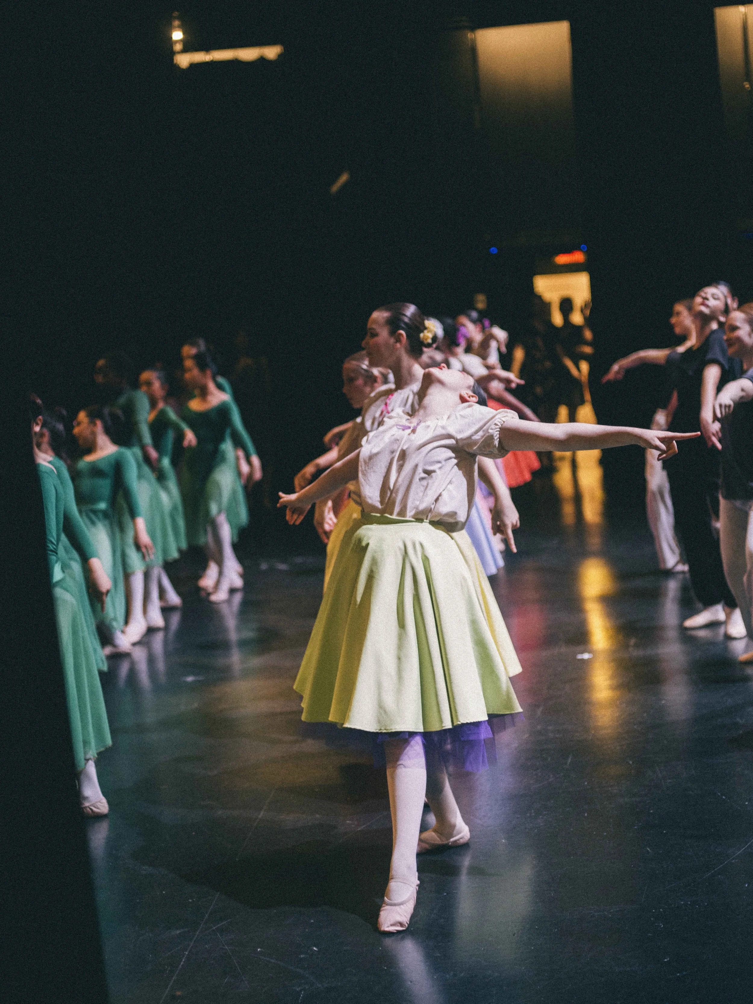 A group of ballet dancers rehearsing backstage, with one dancer in a yellow skirt and beige top in the foreground, all stretching and practicing ballet moves.