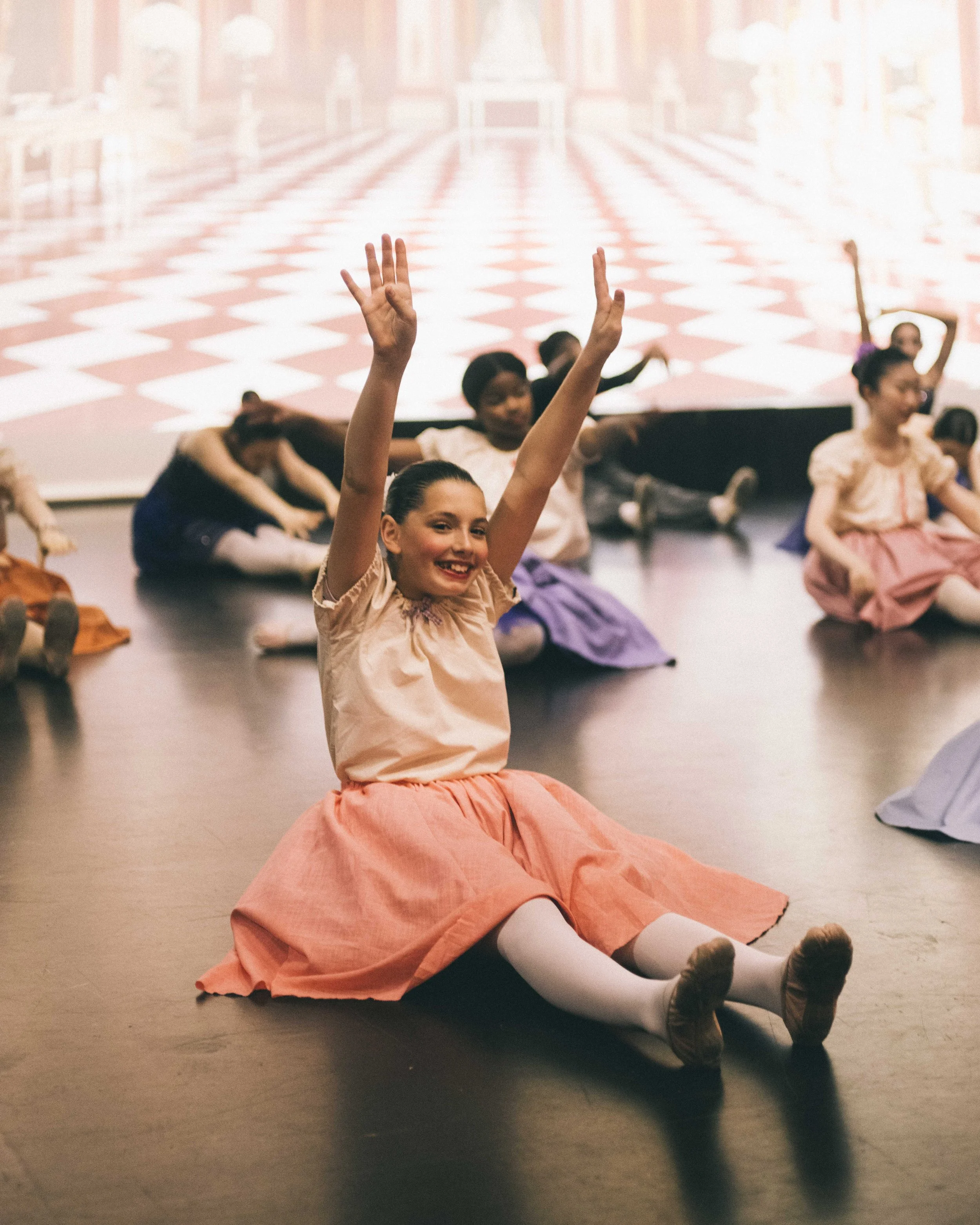 Young girl sitting cross-legged on the floor with arms raised and smiling in a group dance class, with other children sitting nearby on the floor.