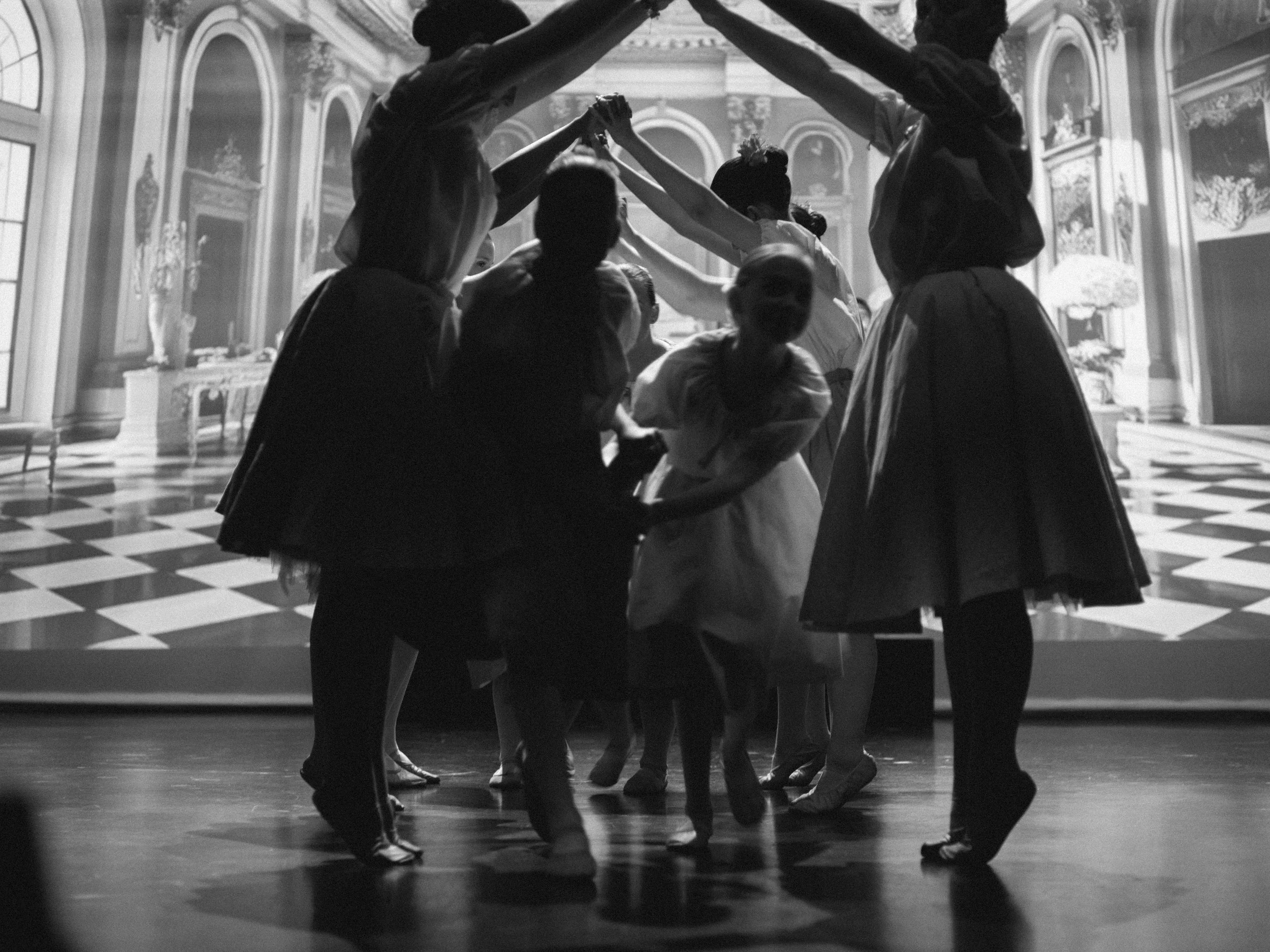 A group of children and adults performing a dance or theatrical act on stage with a backdrop of an ornate, classical interior scene in black and white.