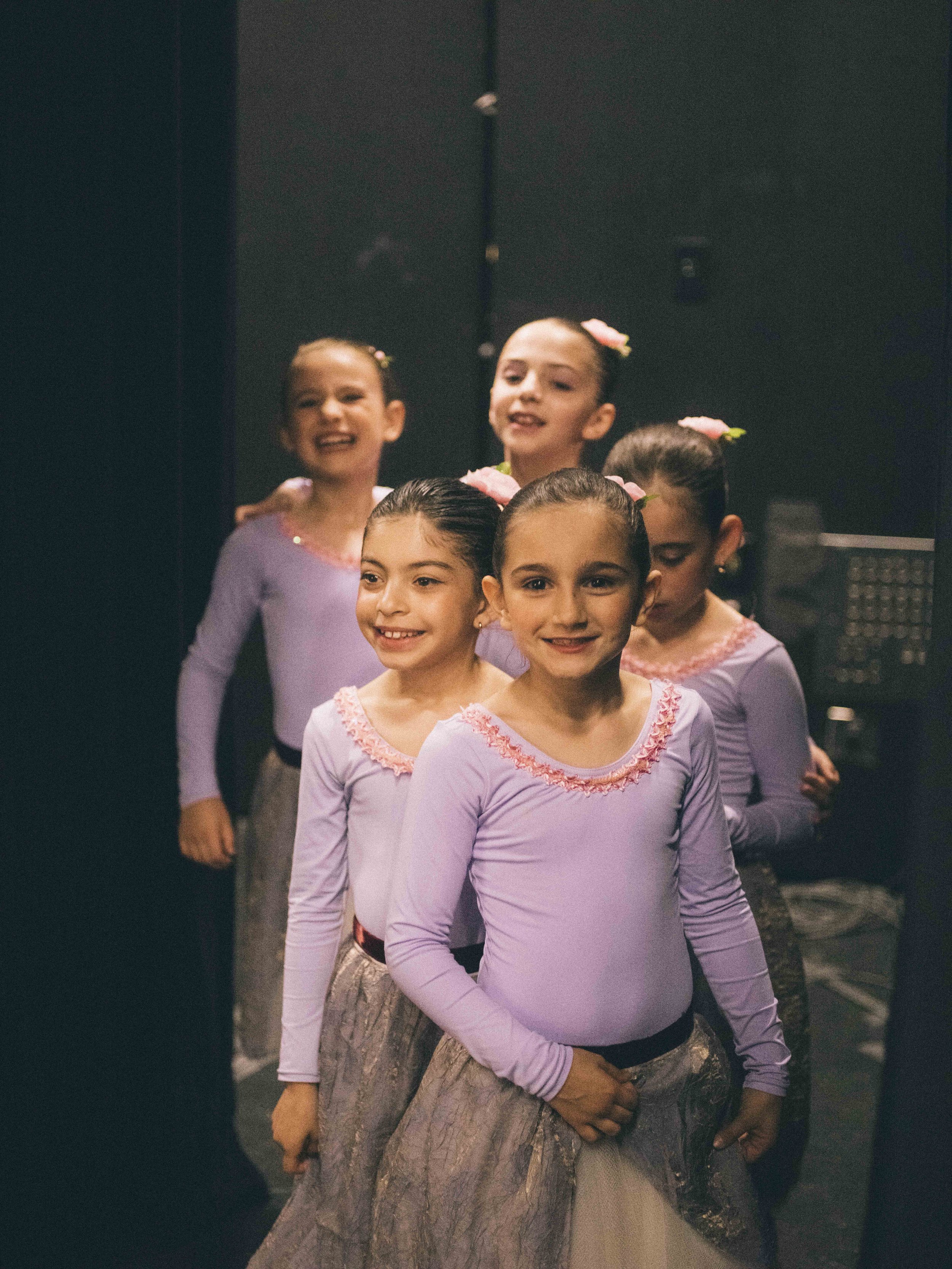 Five young girls in lavender ballet costumes backstage, smiling and preparing for a dance performance.