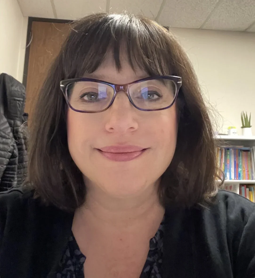 A woman with glasses and dark brown hair taking a selfie in an office setting, with bookshelves and a potted plant in the background.